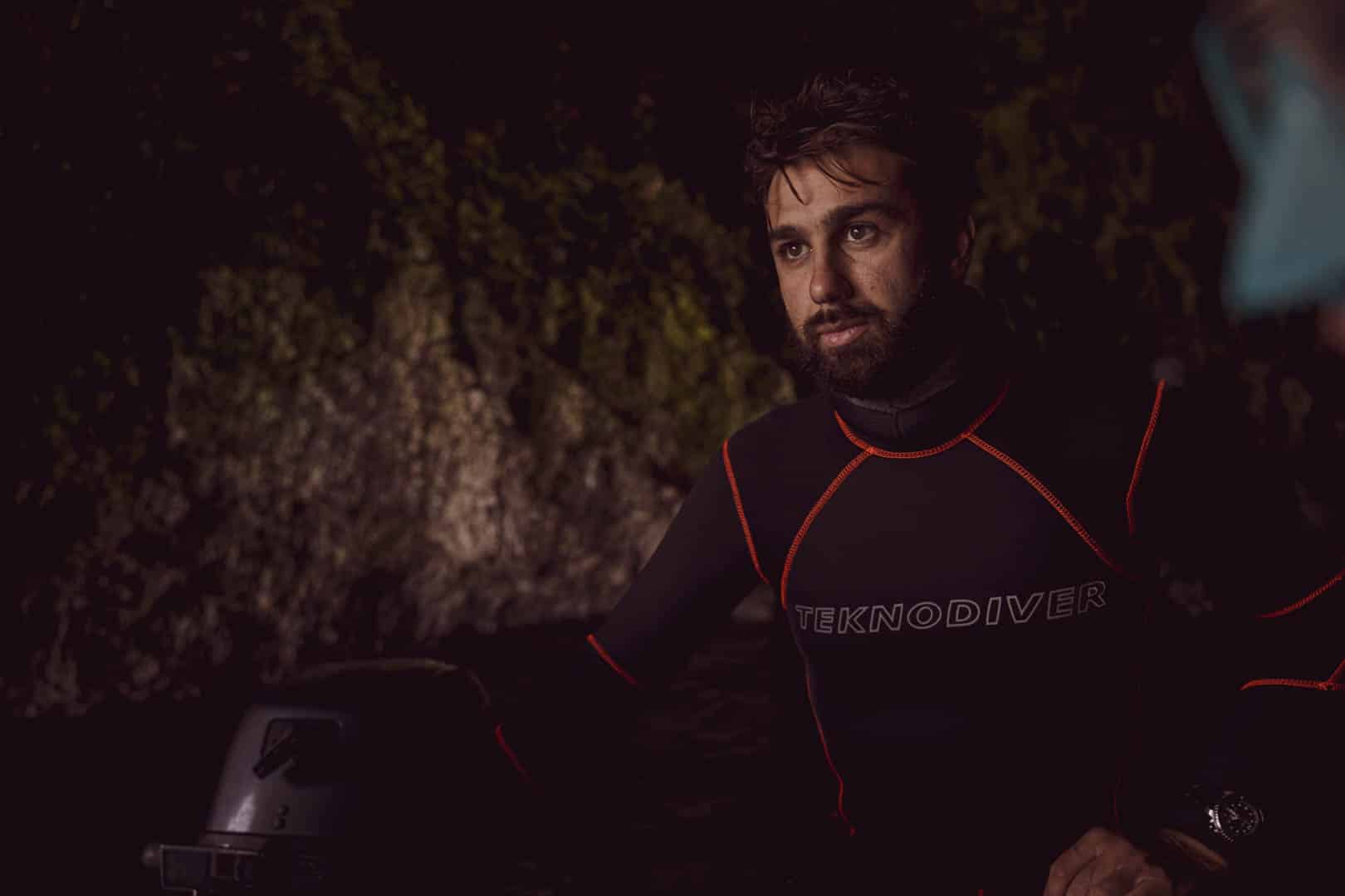 Ein Mann in einem dunklen Neoprenanzug mit der Aufschrift "TEKNODIVER" steht in einer schwach beleuchteten Höhle und schaut konzentriert. Im Hintergrund sind moosbewachsene Felswände zu sehen. © Fotografie Tomas Rodriguez
