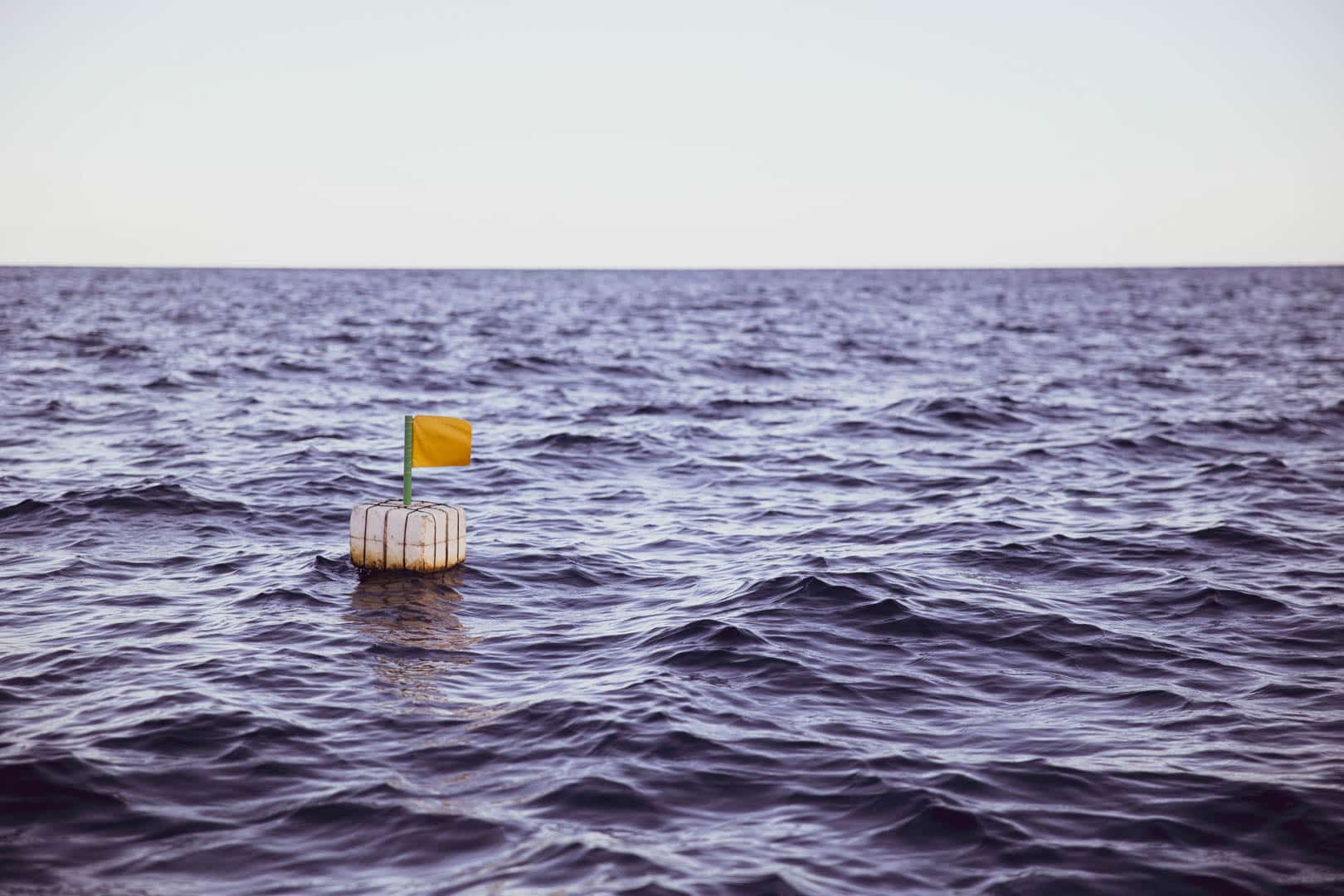 Eine kleine weiße Boje mit einer gelben Flagge schwimmt allein auf dunklem, kräuselndem Wasser unter einem blassen Himmel. © Fotografie Tomas Rodriguez