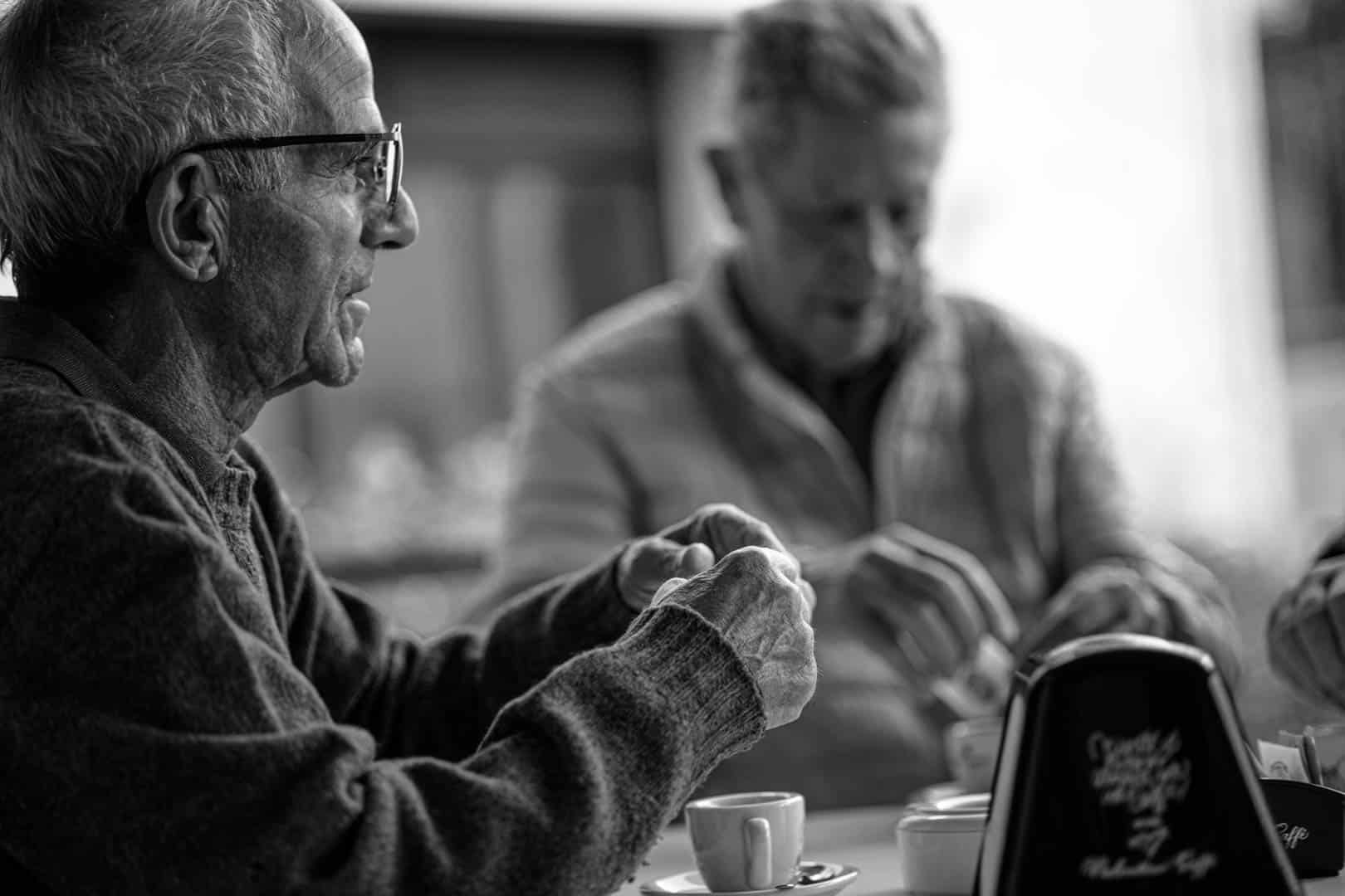 Zwei ältere Männer sitzen an einem Tisch in einem Café und bereiten ihren Kaffee zu. Ein Mann mit Brille und Pullover steht im Mittelpunkt, während der andere unscharf im Hintergrund zu sehen ist. Beide wirken entspannt und in ein Gespräch vertieft. © Fotografie Tomas Rodriguez