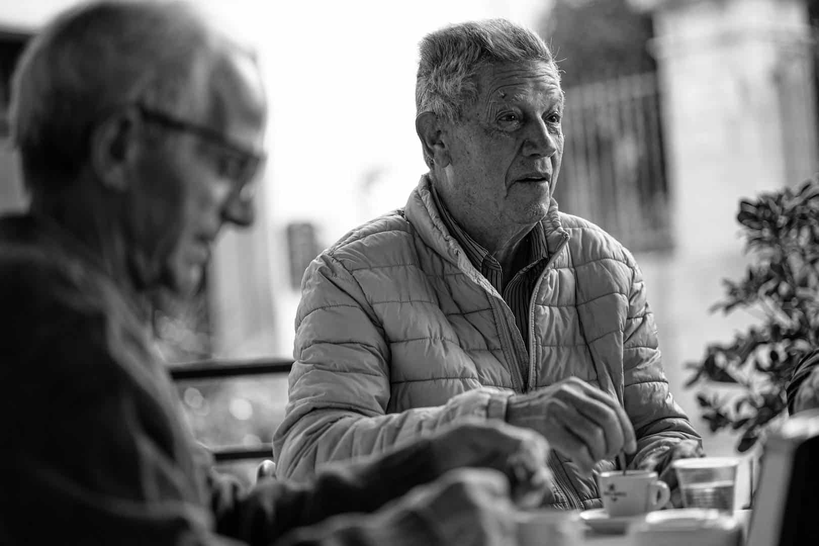 Zwei ältere Männer sitzen an einem Tisch im Freien und trinken Kaffee. Der eine Mann, der im Fokus steht, trägt eine helle Steppjacke und blickt zur Seite. Der andere ist unscharf im Vordergrund. Die Szene ist in schwarz-weiß gehalten. © Fotografie Tomas Rodriguez