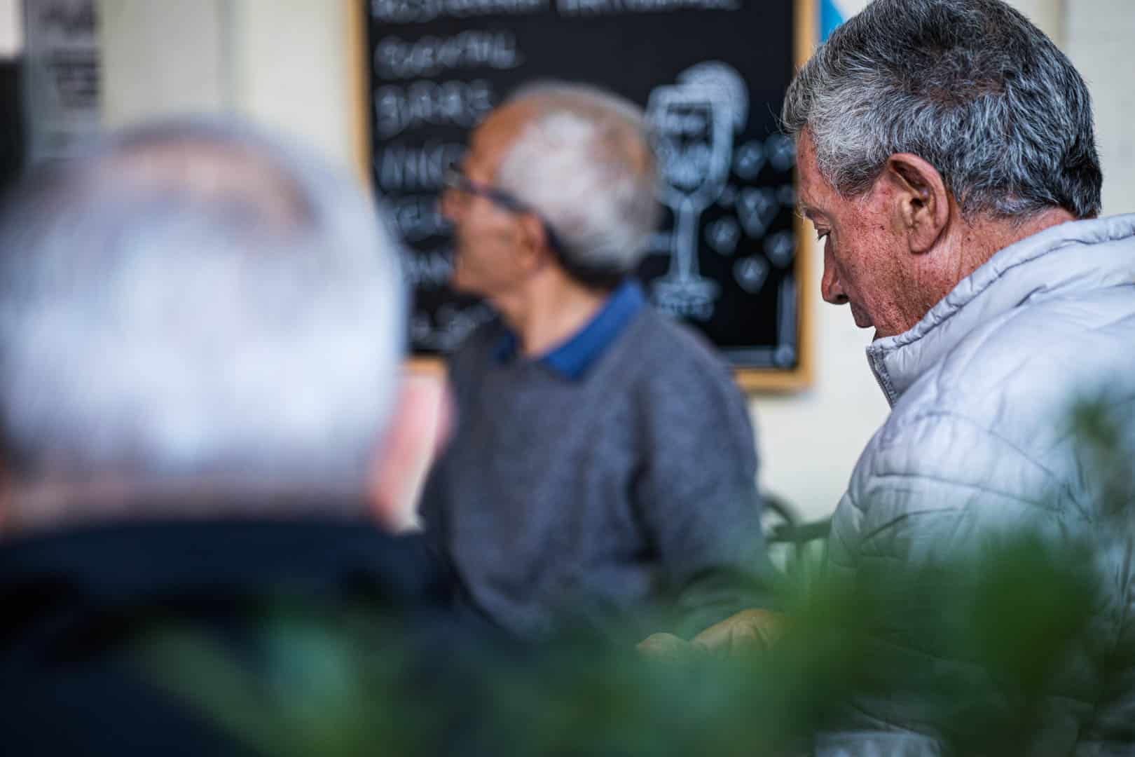 Drei ältere Männer, zwei mit grauem Haar im Vordergrund und einer im Hintergrund, sitzen in einem Innenraum in der Nähe einer Speisekarte mit Kreidetafel. Der Fokus liegt auf dem Mann mit der hellen Jacke auf der rechten Seite, während die anderen unscharf sind. © Fotografie Tomas Rodriguez