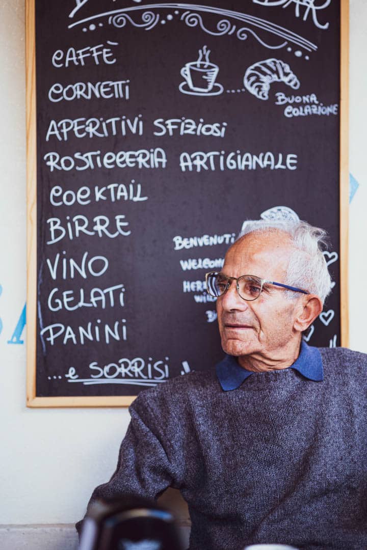 Ein älterer Mann mit Brille sitzt vor einer mit Kreide geschriebenen Speisekarte in italienischer Sprache, auf der Dinge wie caffè, cornetti, birre, vino, gelati und panini aufgeführt sind. Er schaut zur Seite und wirkt nachdenklich. © Fotografie Tomas Rodriguez