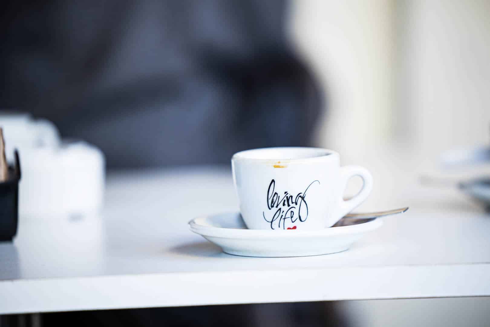 Eine weiße Espressotasse mit einer Untertasse und einem Löffel steht auf einem weißen Tisch. Die Tasse ist in schwarzer Schreibschrift beschriftet. Der Hintergrund ist unscharf. © Fotografie Tomas Rodriguez