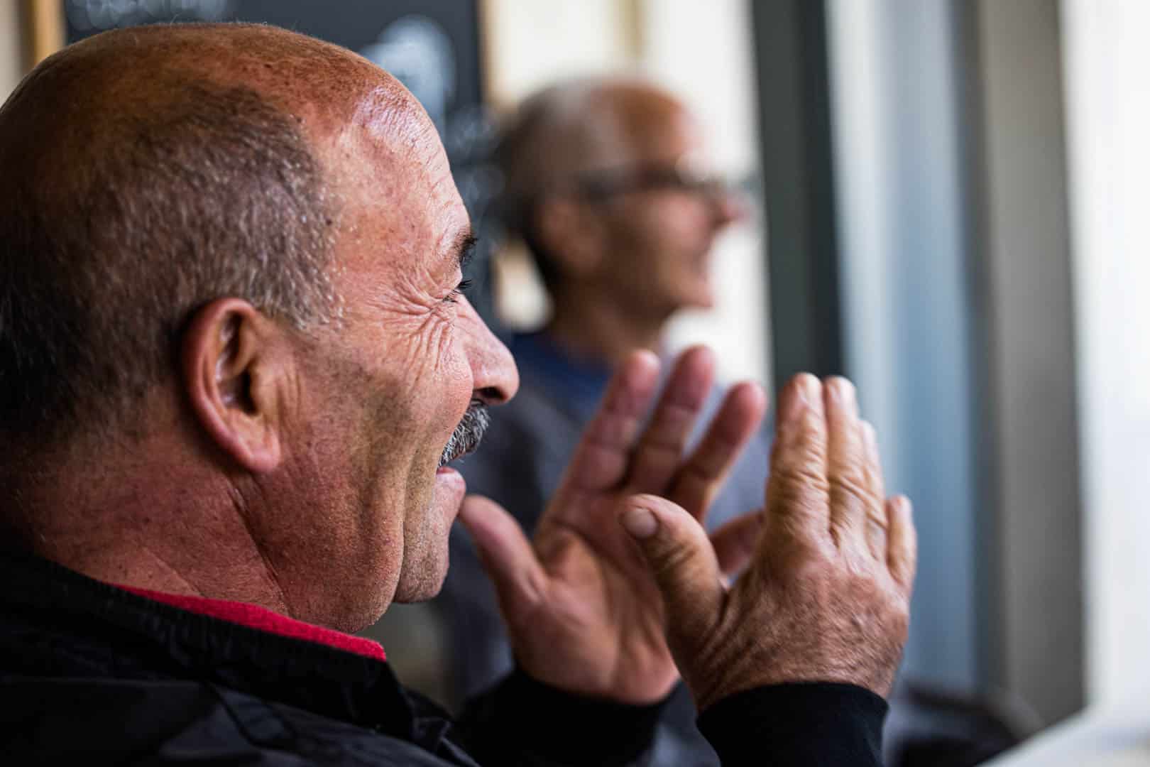 Nahaufnahme eines älteren Mannes mit Schnurrbart, der lächelt und klatscht, während ein anderer älterer Mann im Hintergrund verschwommen zu erkennen ist. Beide scheinen sich in einem Haus in der Nähe eines Fensters zu befinden. © Fotografie Tomas Rodriguez