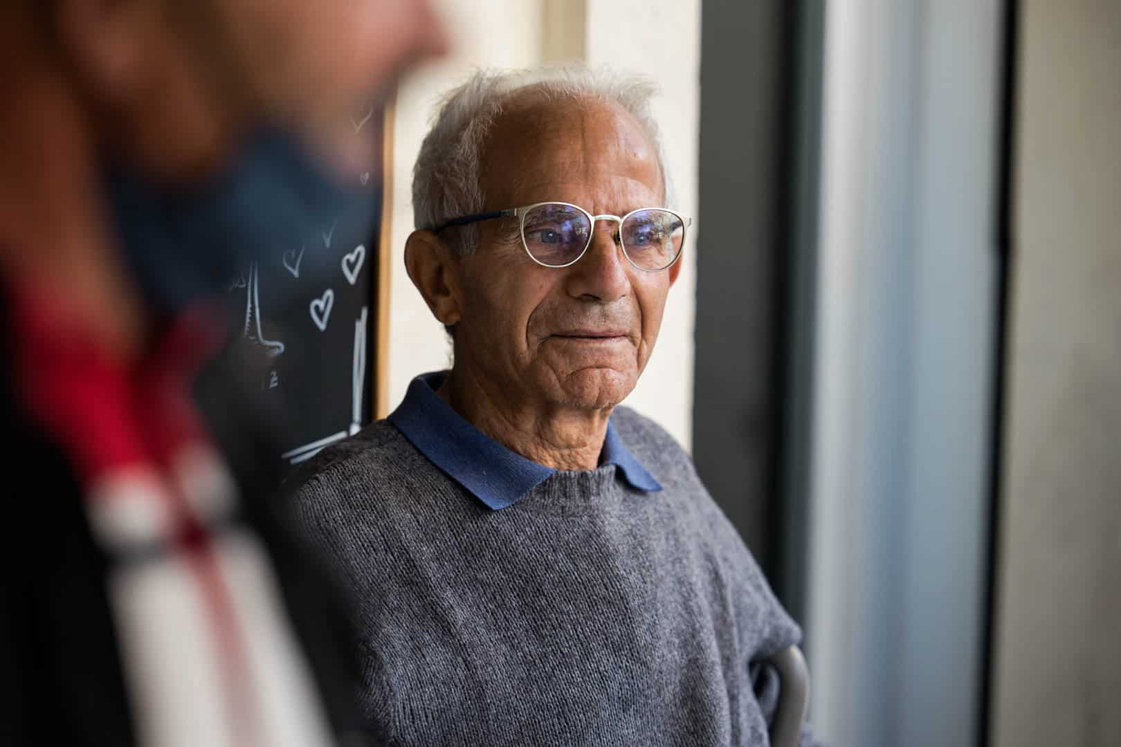 Ein älterer Mann mit weißem Haar und Brille, der einen grauen Pullover mit blauem Kragen trägt, sitzt in einem Haus und schaut nachdenklich nach vorn. Eine weitere Person ist unscharf im Vordergrund zu erkennen. © Fotografie Tomas Rodriguez