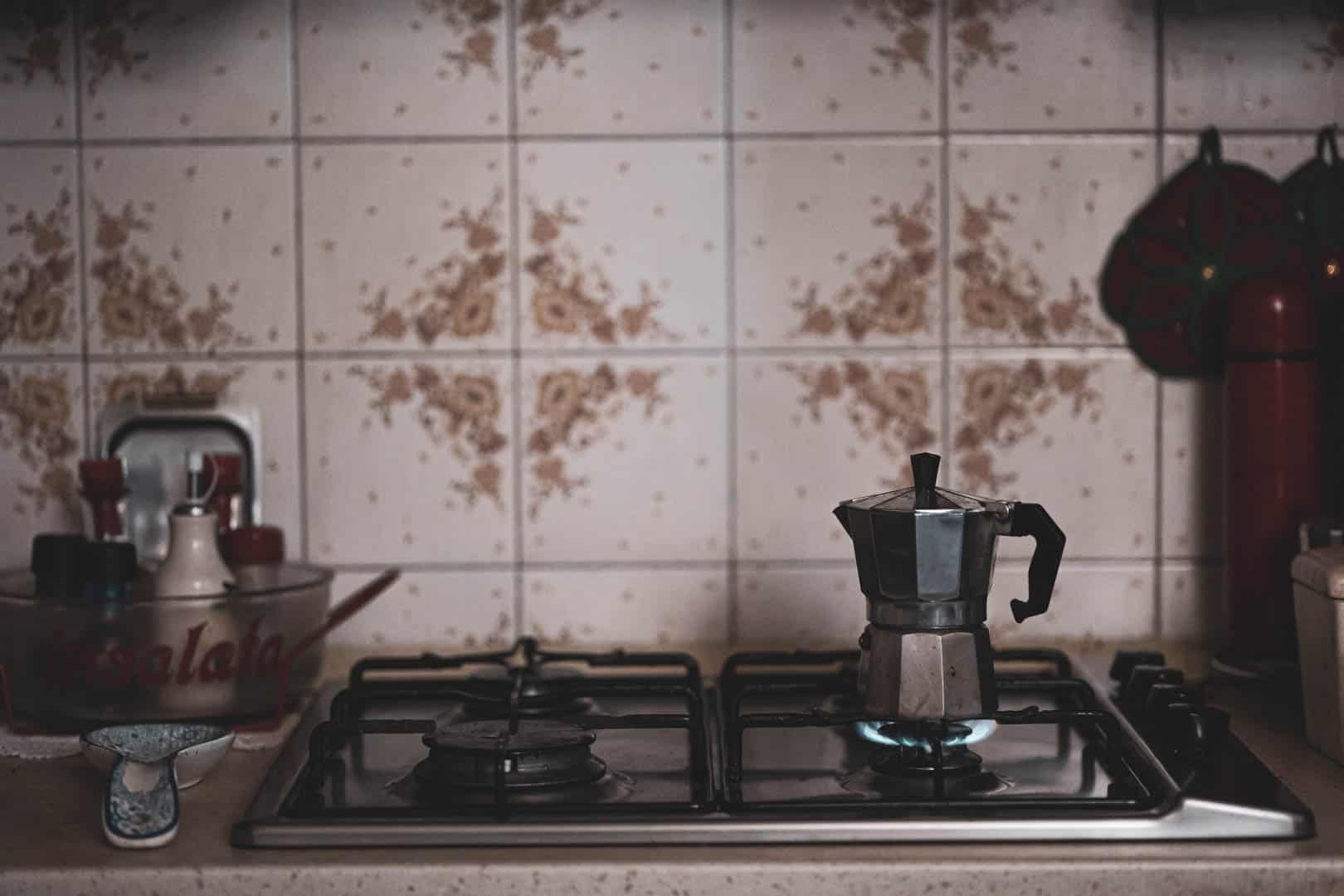 Auf einem Gasherd mit blauer Flamme in einer Vintage-Küche brüht eine Moka-Kanne. Im Hintergrund sind floral gemusterte Kacheln und verschiedene Küchenutensilien auf dem Tresen zu sehen. © Fotografie Tomas Rodriguez