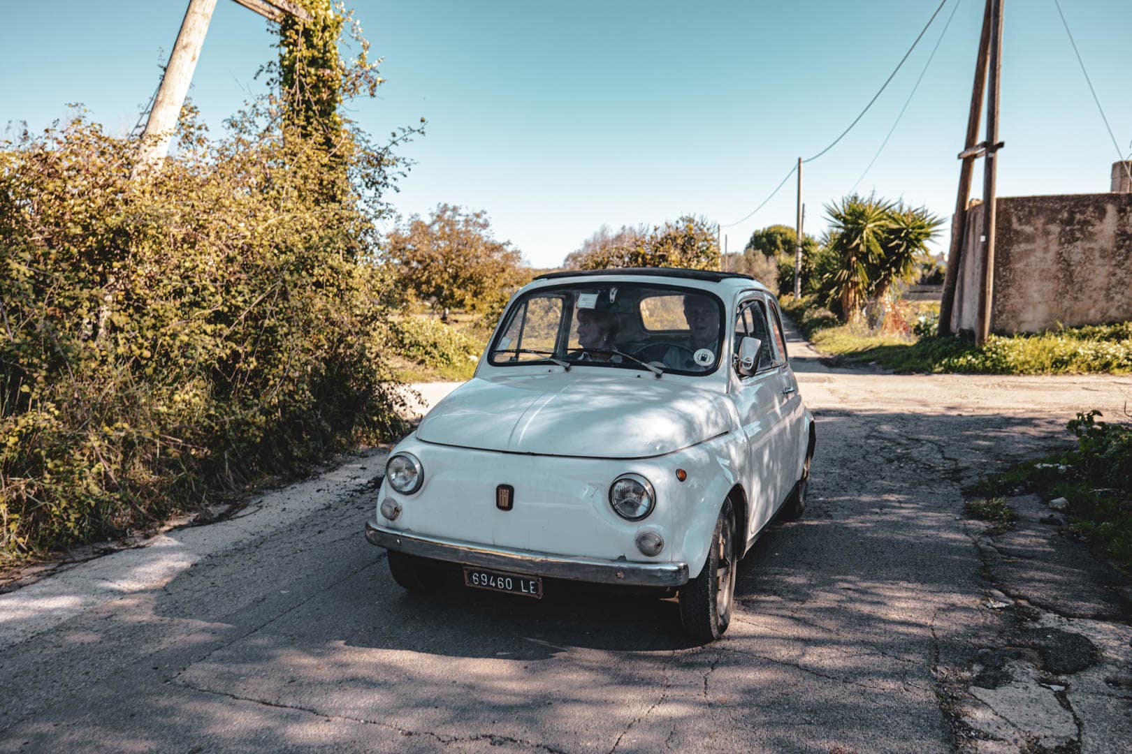 Ein klassischer weißer Fiat 500 ist auf einer sonnenbeschienenen, rissigen Landstraße geparkt, umgeben von überwucherten Büschen und Bäumen unter einem klaren blauen Himmel. © Fotografie Tomas Rodriguez