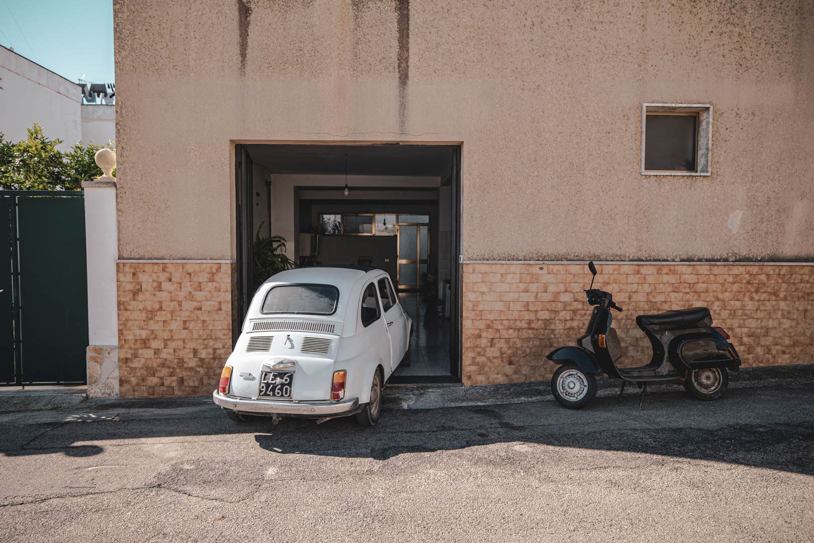 Ein kleiner weißer Oldtimer ist teilweise in einer Garage eines beigen Gebäudes geparkt, neben einem schwarzen Motorroller, der auf der Straße geparkt ist. Das Garagentor ist geöffnet und gibt den Blick auf einen Flur im Inneren des Gebäudes frei. © Fotografie Tomas Rodriguez