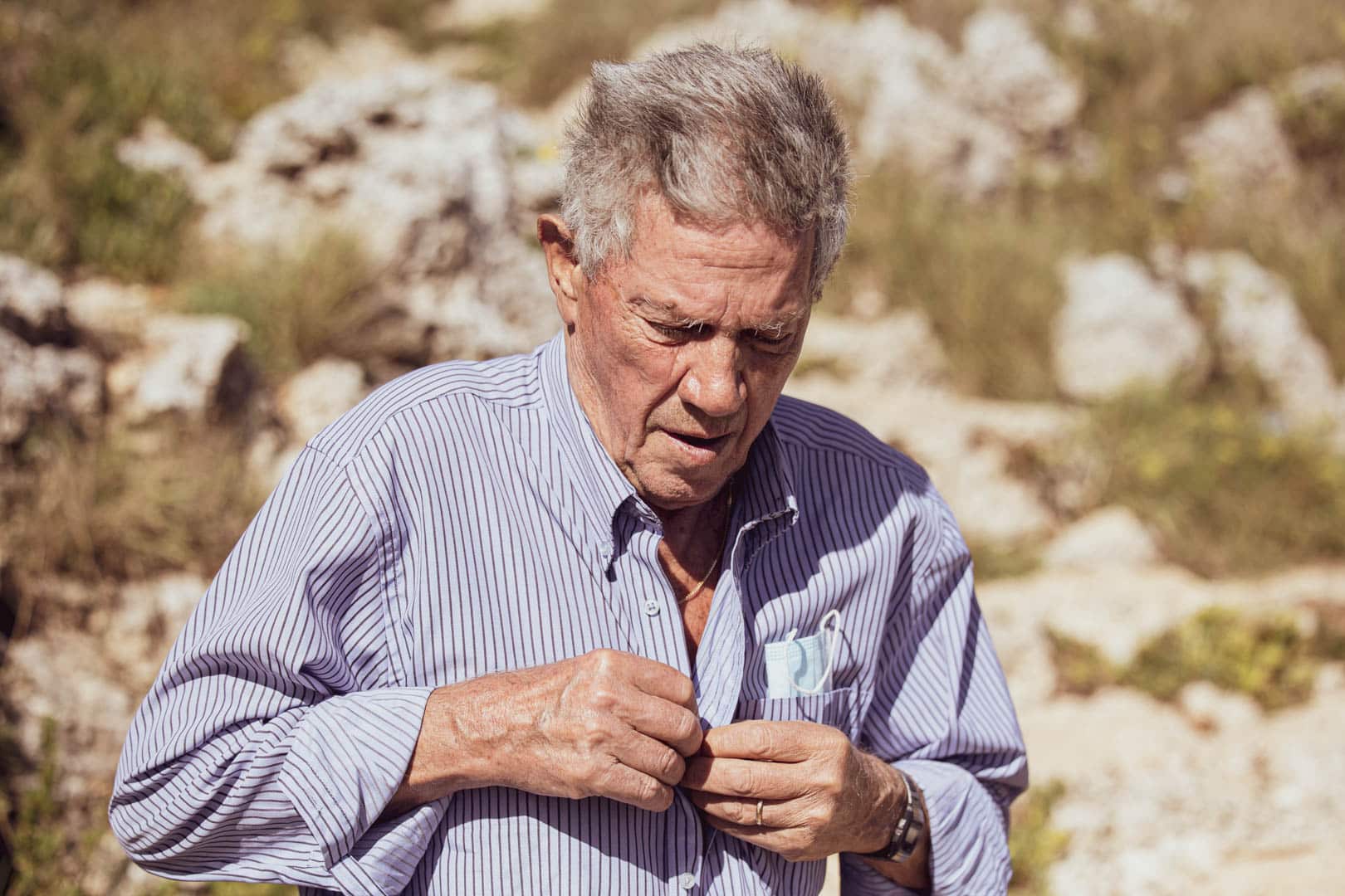 Ein älterer Mann, der ein blau-weiß gestreiftes Hemd trägt, knöpft es im Freien zu. Er schaut mit konzentriertem Blick nach unten, und im unscharfen Hintergrund sind Felsen und Gras zu sehen. © Fotografie Tomas Rodriguez