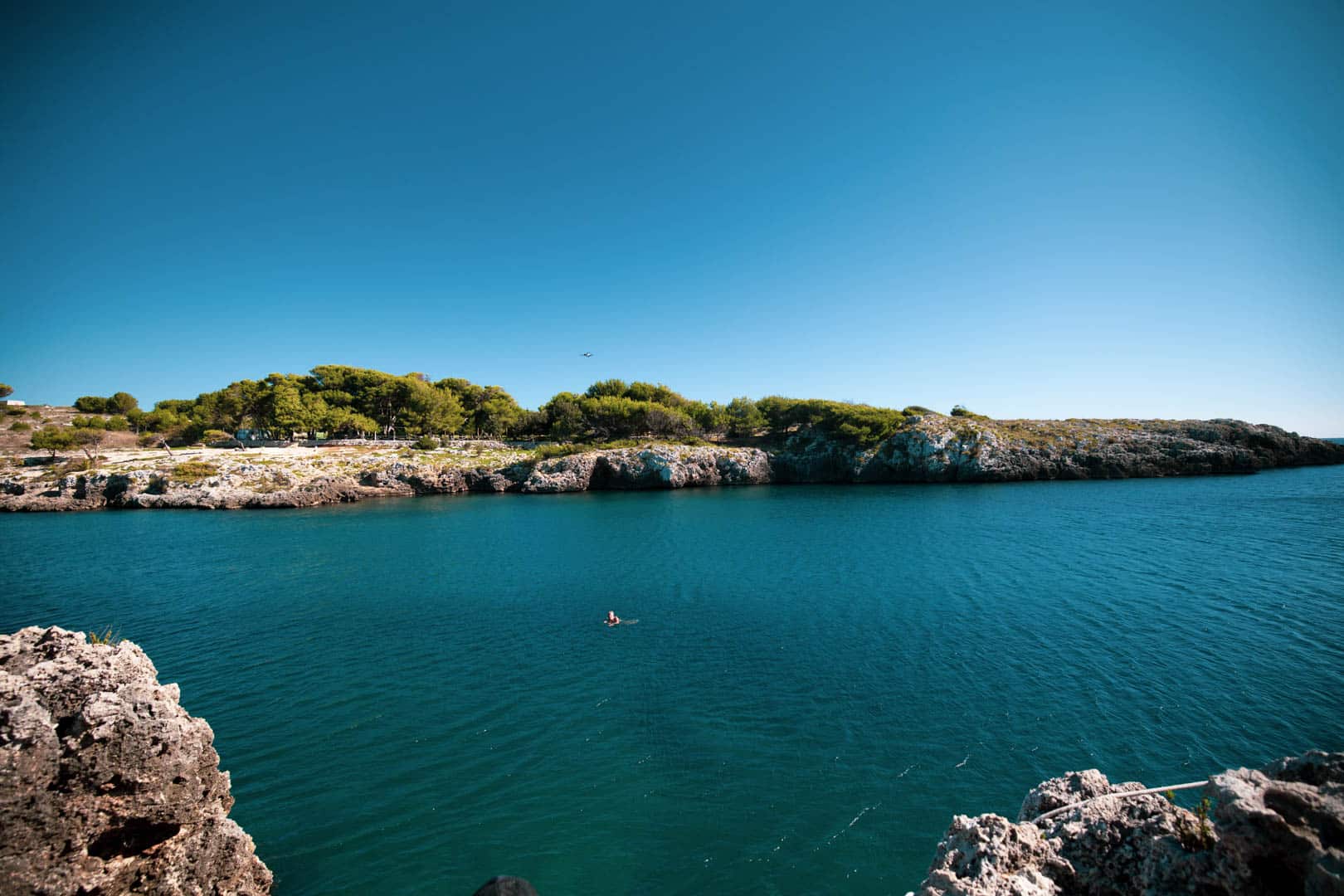 Eine Person schwimmt allein im klaren blauen Wasser in der Nähe von Felsen, mit einem baumbewachsenen Ufer und einem strahlend blauen Himmel im Hintergrund. © Fotografie Tomas Rodriguez