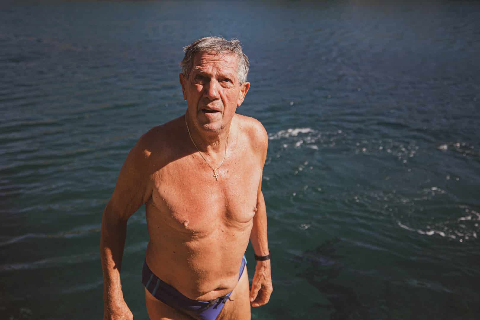 Ein älterer Mann mit grauem Haar steht hüfttief in einem Gewässer und trägt einen Badeslip. Er blickt leicht nach oben und hat einen neutralen Blick. Das Sonnenlicht beleuchtet seine nasse Haut und das plätschernde blaue Wasser hinter ihm. © Fotografie Tomas Rodriguez