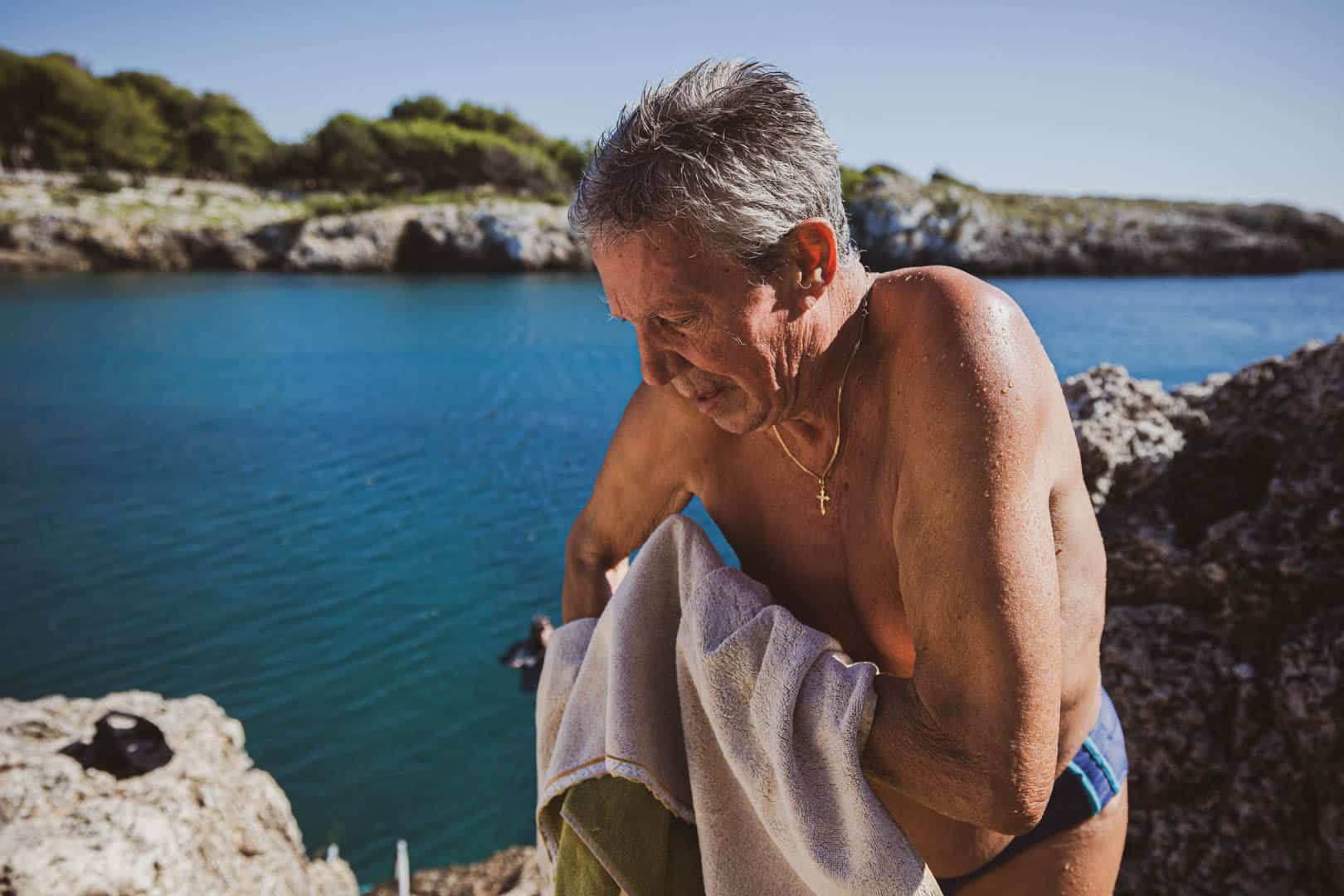 Ein älterer Mann mit nassen Haaren steht auf einem felsigen Gelände am Meer und trocknet sich mit einem Handtuch ab. Er trägt eine Badehose und eine Halskette mit einem Kreuz. Im Hintergrund sind blaues Wasser und felsige Klippen zu sehen. © Fotografie Tomas Rodriguez