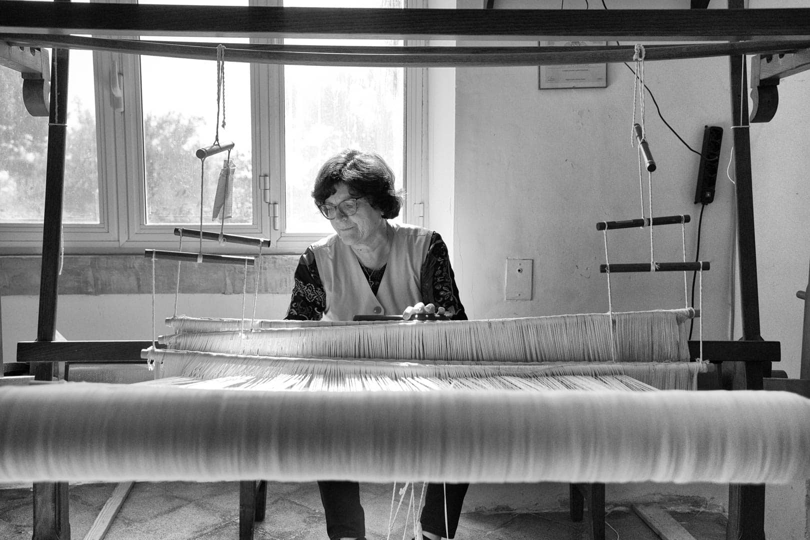 Eine Frau sitzt an einem Webstuhl und webt in einem sonnendurchfluteten Raum mit einem Fenster im Hintergrund von Hand Stoff. Das Foto ist in Schwarz-Weiß gehalten und hebt die Fäden und ihren konzentrierten Ausdruck hervor. © Fotografie Tomas Rodriguez