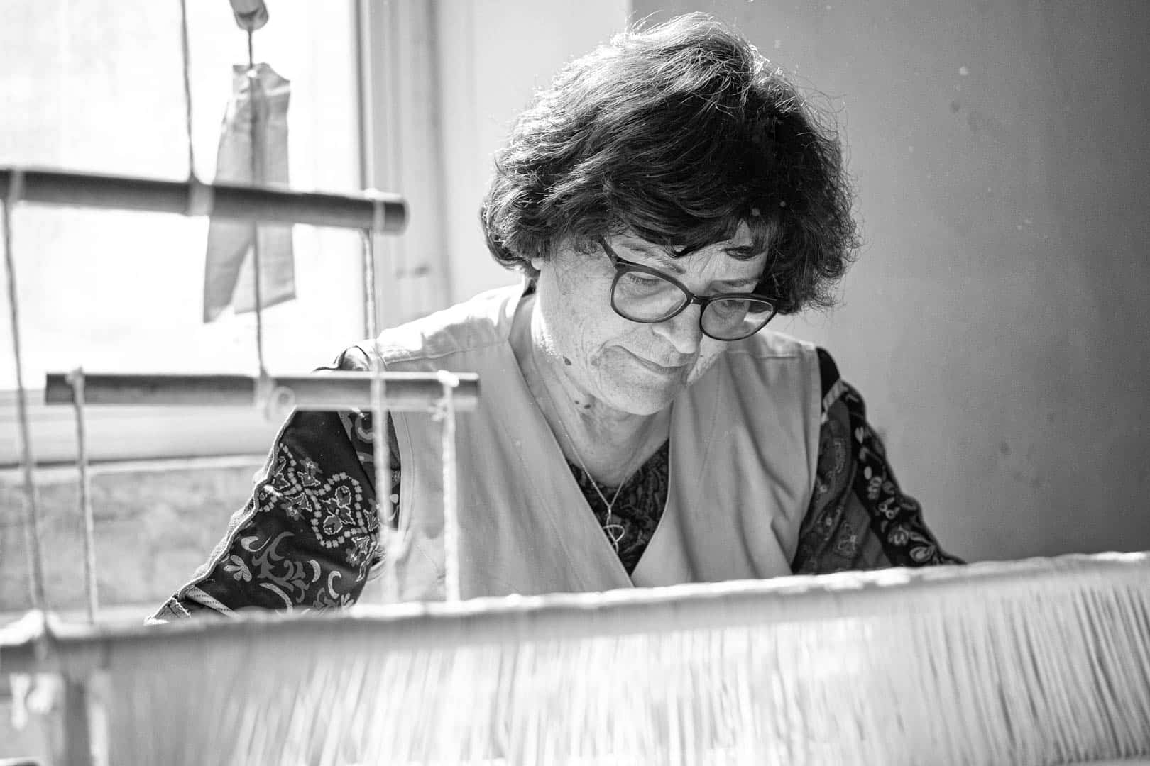 Schwarz-Weiß-Foto einer älteren Frau mit Brille und lockigem Haar, die konzentriert an einem Webstuhl in der Nähe eines Fensters arbeitet und bei natürlichem Licht Stoff webt. © Fotografie Tomas Rodriguez