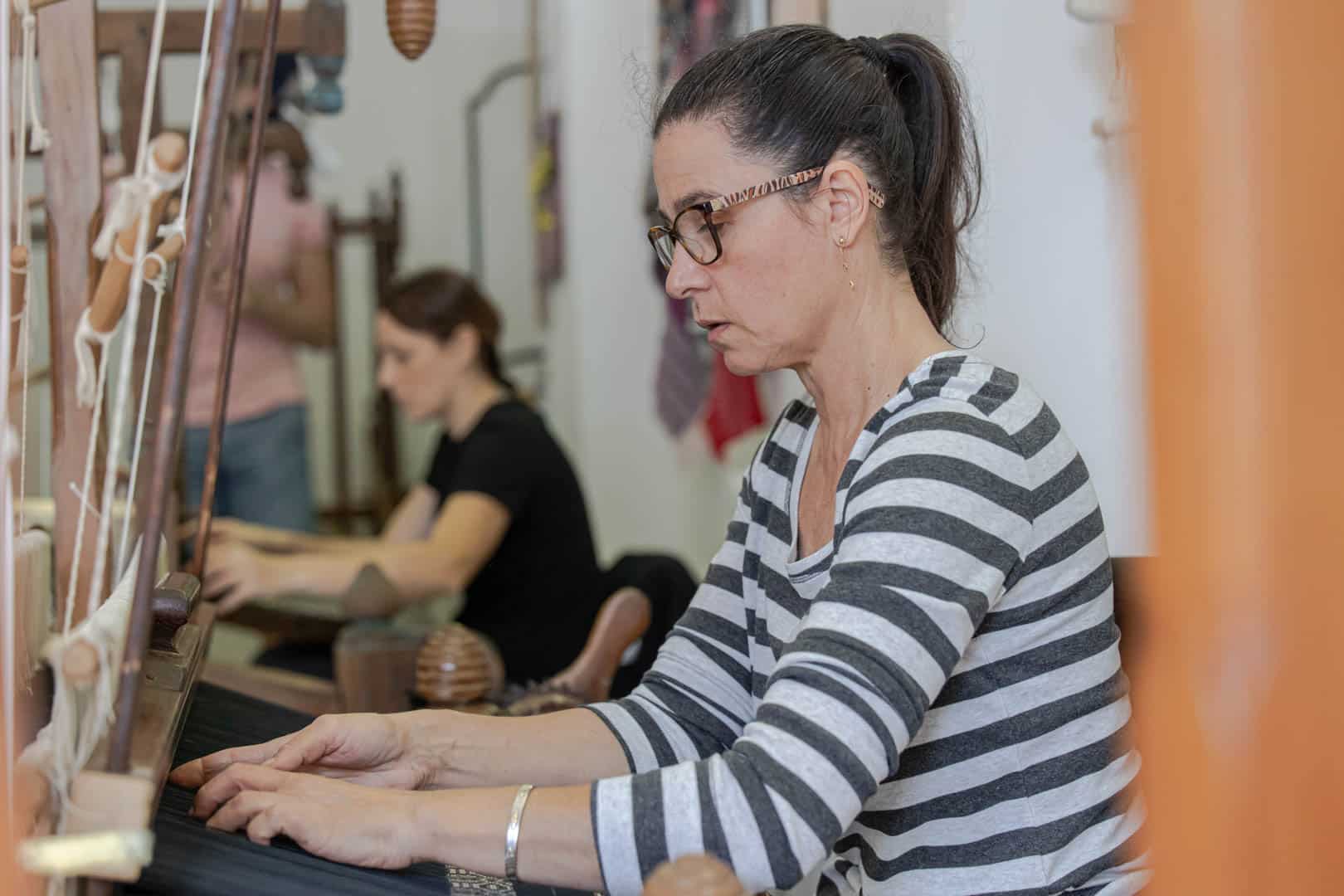 Eine Frau mit Brille und gestreiftem Hemd webt Stoff auf einem Webstuhl und konzentriert sich auf ihre Arbeit. Eine andere Person webt im Hintergrund in einer Werkstatt. © Fotografie Tomas Rodriguez
