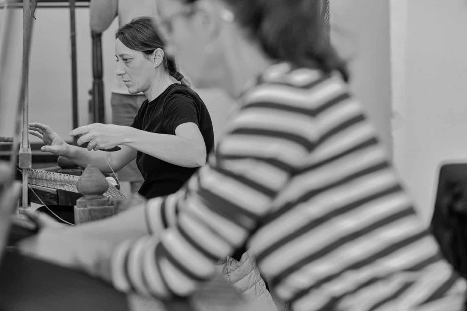 Zwei Frauen, die nebeneinander arbeiten, eine im Fokus, die ein dunkles Hemd trägt und webt, die andere im Vordergrund, die ein gestreiftes Hemd trägt, unscharf. Die Umgebung scheint eine Werkstatt oder ein Atelier zu sein. Schwarz-Weiß-Foto. © Fotografie Tomas Rodriguez