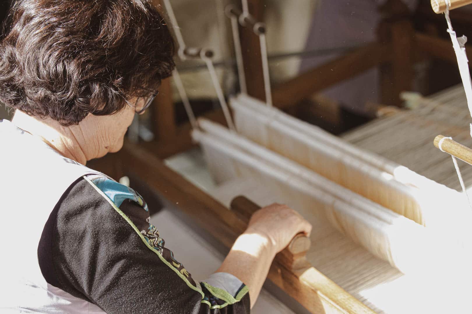 Eine Frau mit kurzen dunklen Haaren bedient einen traditionellen Holzwebstuhl und webt im Sonnenlicht weiße Fäden. Ihre Hände führen den Webmechanismus, während sie sich auf ihre Arbeit konzentriert. © Fotografie Tomas Rodriguez