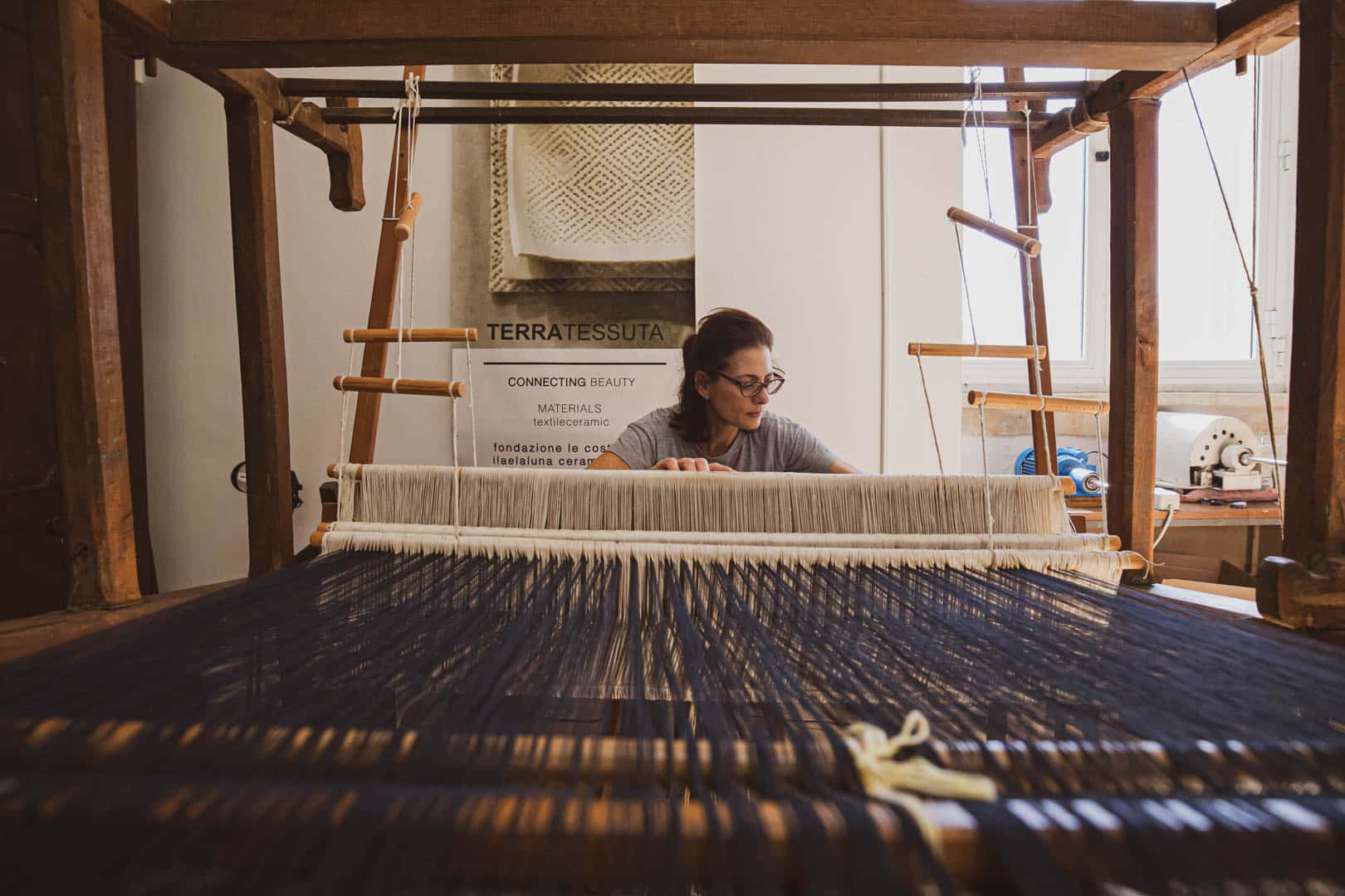 Eine Frau sitzt hinter einem großen hölzernen Webstuhl und webt dunkle und helle Fäden in einem hellen Raum mit einem Fenster und Textilkunst an der Wand im Hintergrund. © Fotografie Tomas Rodriguez
