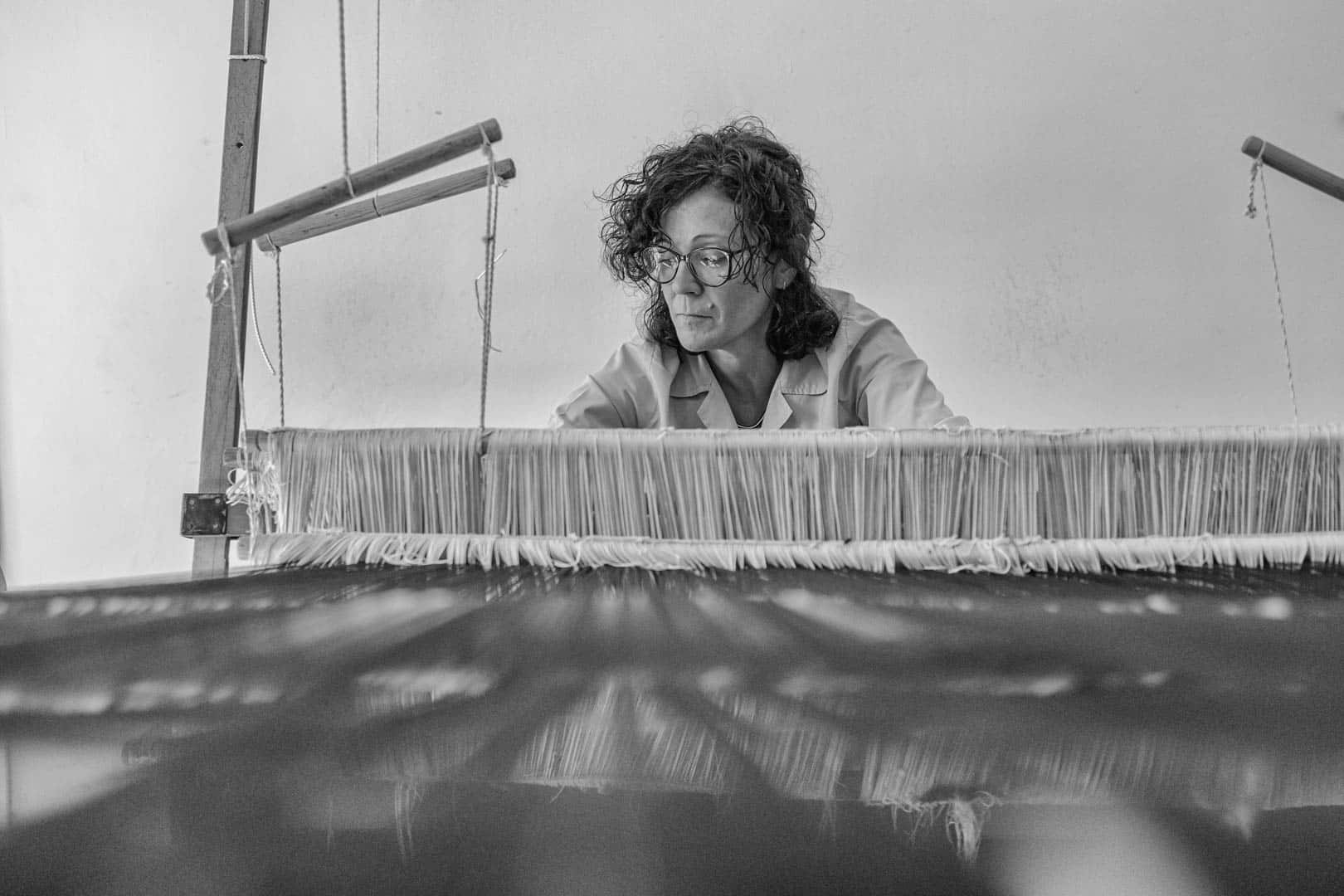 Eine Frau mit lockigem Haar und Brille arbeitet konzentriert an einem großen Webstuhl und webt Textilien. Das Bild ist in Schwarz-Weiß gehalten und unterstreicht die konzentrierte Atmosphäre im Arbeitsbereich. © Fotografie Tomas Rodriguez