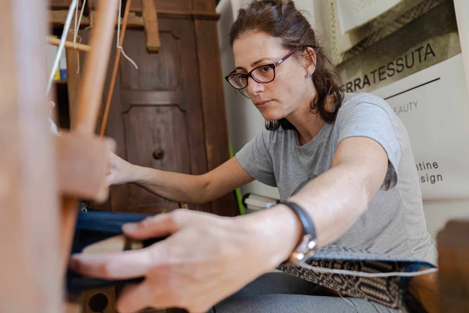 Eine Frau, die eine Brille und ein graues Hemd trägt, konzentriert sich auf das Weben von Stoffen auf einem Webstuhl in einem Innenbereich. Sie positioniert die Fäden sorgfältig mit beiden Händen. © Fotografie Tomas Rodriguez