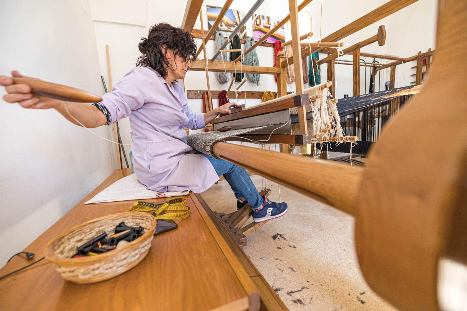Eine Frau bedient einen traditionellen Holzwebstuhl und webt in einem hellen Raum Stoff. Sie konzentriert sich auf ihre Arbeit, während die Webwerkzeuge und Garnspulen in einem Korb in der Nähe liegen. © Fotografie Tomas Rodriguez