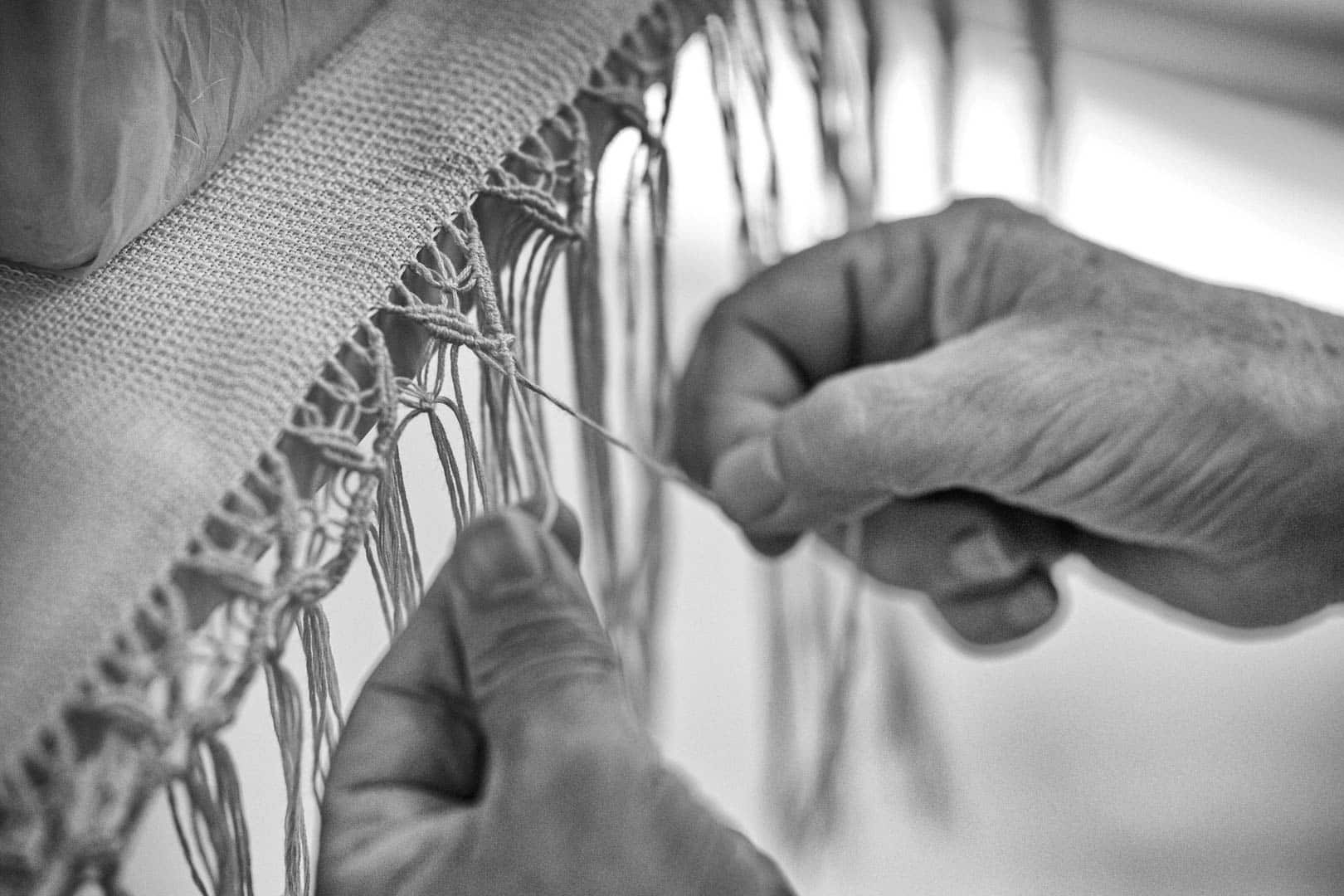 Nahaufnahme von zwei Händen, die sorgfältig Fransen auf ein Stück Stoff weben oder knüpfen, wobei die detaillierte Handwerkskunst hervorgehoben wird. Das Bild ist in schwarz-weiß gehalten. © Fotografie Tomas Rodriguez