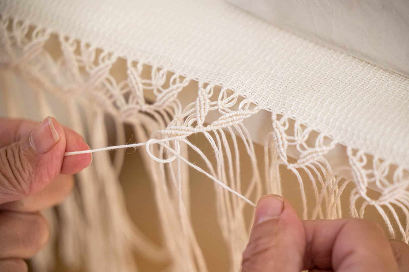 Nahaufnahme von Händen, die mit einem cremefarbenen Faden Knoten weben oder knüpfen und dabei ein dekoratives Makramee- oder Spitzenmuster auf einem Stoffband erzeugen. © Fotografie Tomas Rodriguez
