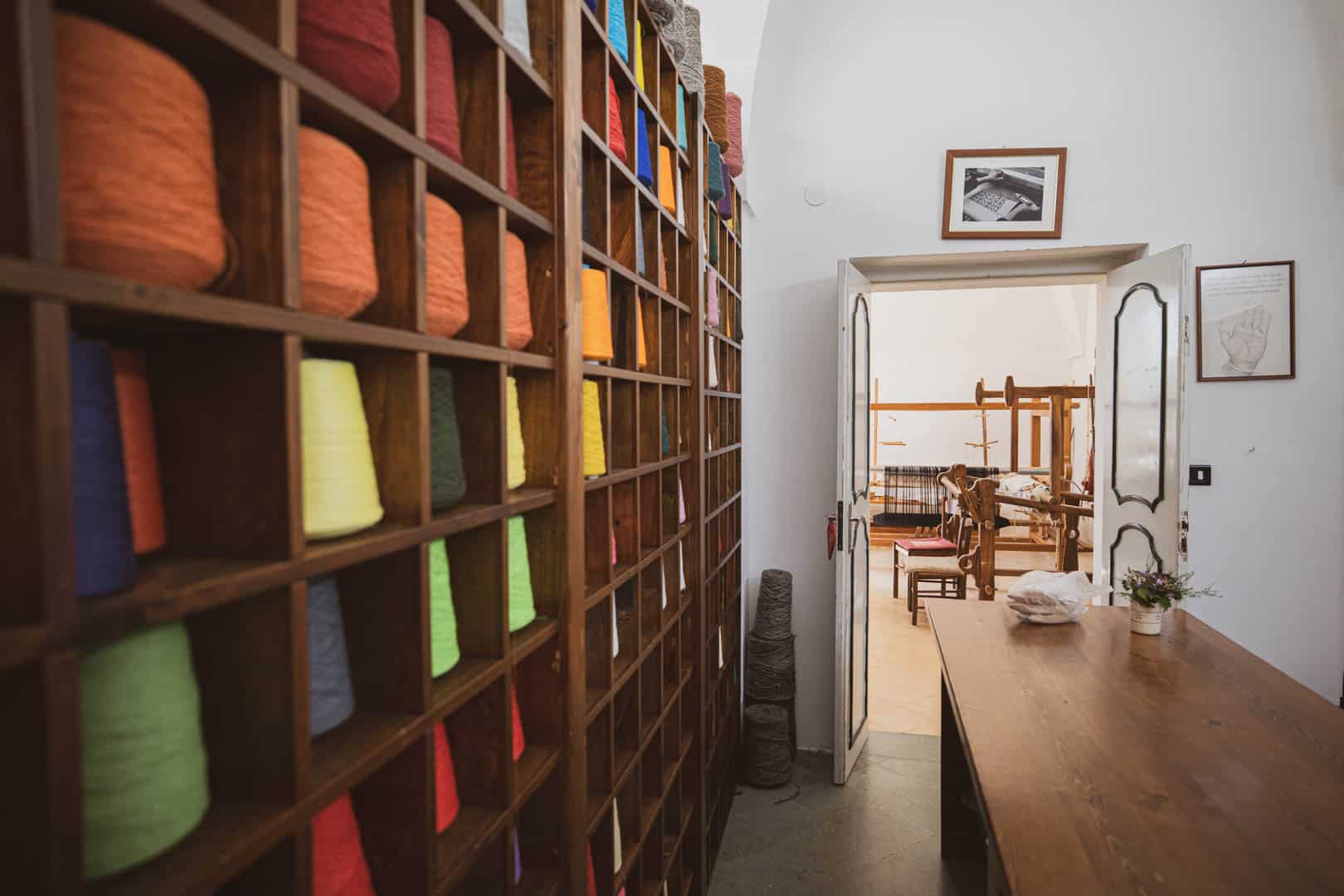 Ein Holzregal mit bunten Garnspulen säumt die linke Wand eines Raumes. Eine offene Tür führt in einen hellen Raum mit einem hölzernen Webstuhl und einem Tisch, die auf eine Textil- oder Webereiwerkstatt hindeuten. © Fotografie Tomas Rodriguez