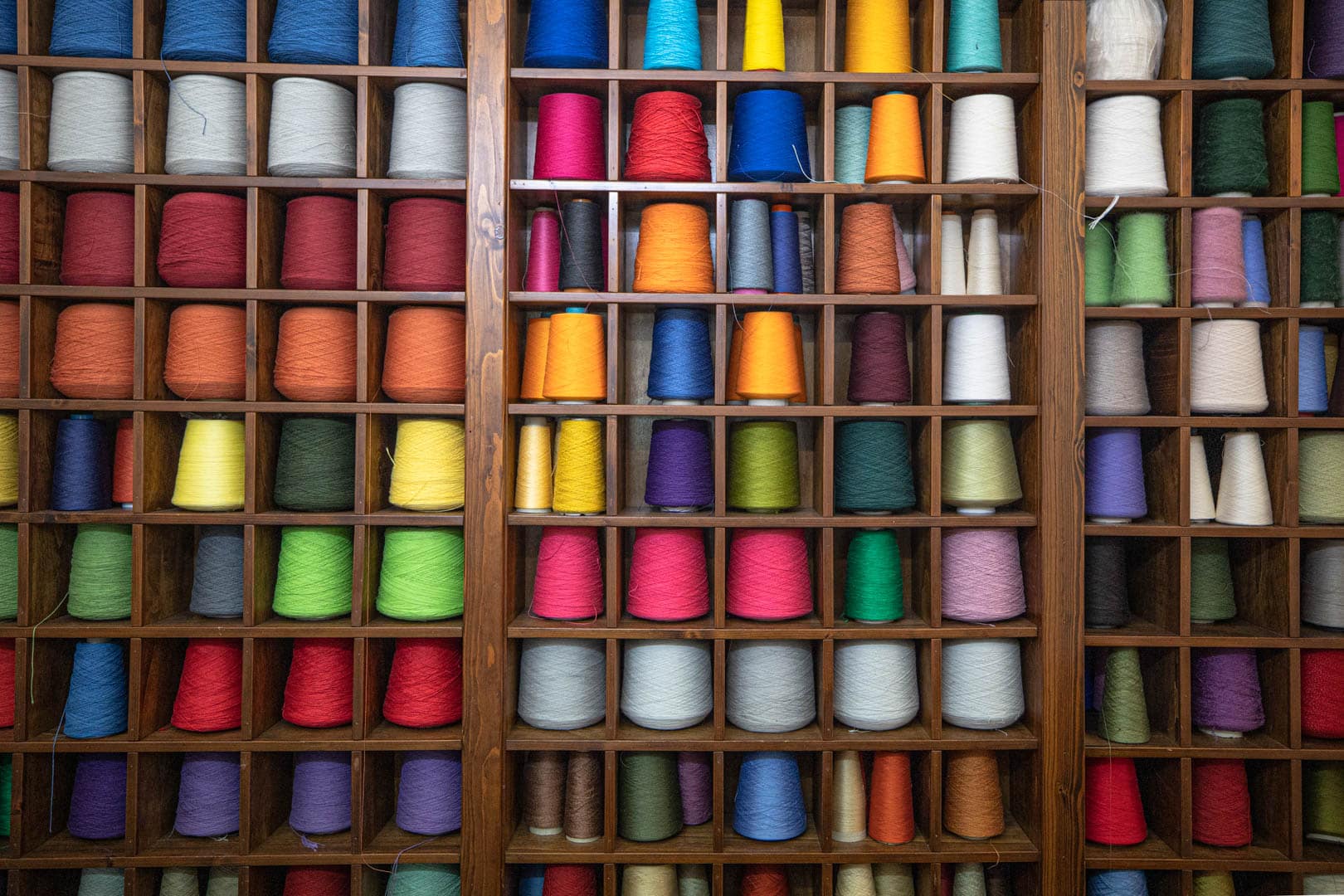 Holzregale, gefüllt mit ordentlich geordneten Reihen bunter Garnspulen in verschiedenen Farbtönen, geordnet nach Farbe und Größe, schaffen eine lebendige und optisch ansprechende Auslage. © Fotografie Tomas Rodriguez
