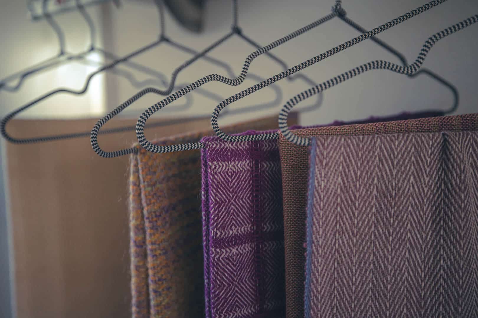 Vier gewebte Schals in verschiedenen Farben und Mustern hängen fein säuberlich auf gestreiften Metallbügeln, die in einer Reihe vor einem neutralen Hintergrund stehen. © Fotografie Tomas Rodriguez