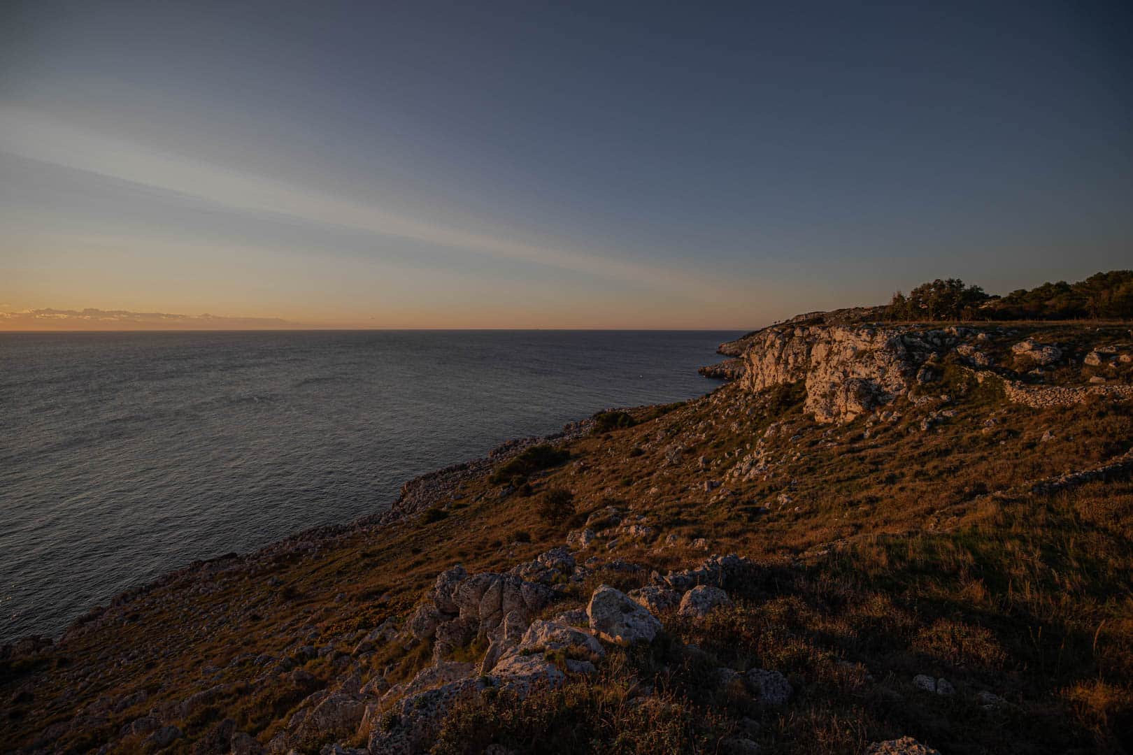 Eine felsige Küste mit zerklüfteten Klippen und spärlicher Vegetation mit Blick auf ein ruhiges Meer bei Sonnenuntergang, wobei der Himmel in der Nähe des Horizonts von warmem Orange zu Blau übergeht. © Fotografie Tomas Rodriguez