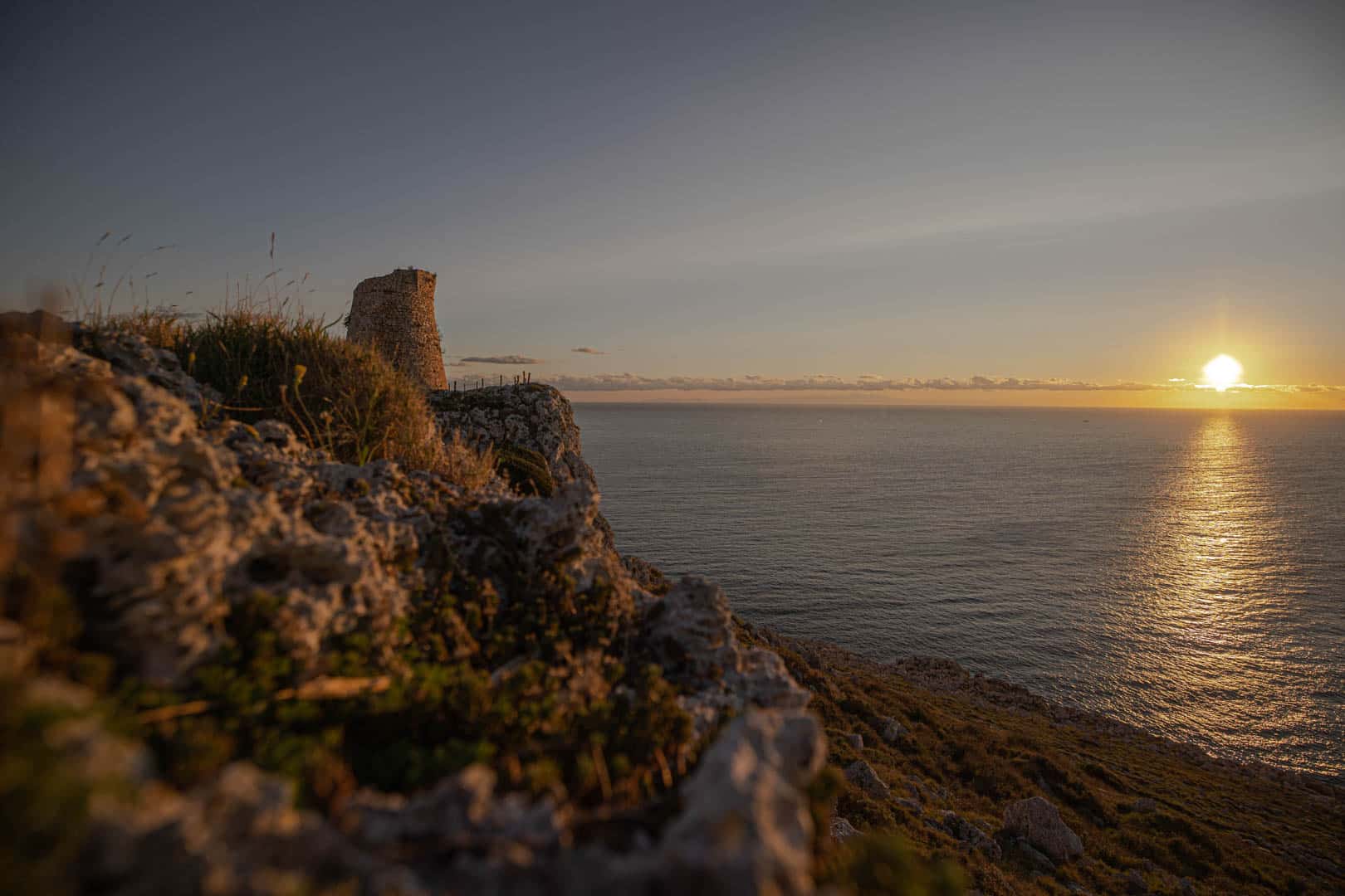 Ein Steinturm steht auf einer felsigen Klippe mit Blick auf ein ruhiges Meer bei Sonnenuntergang, wobei das goldene Sonnenlicht warme Töne über die Landschaft und den Horizont wirft. © Fotografie Tomas Rodriguez