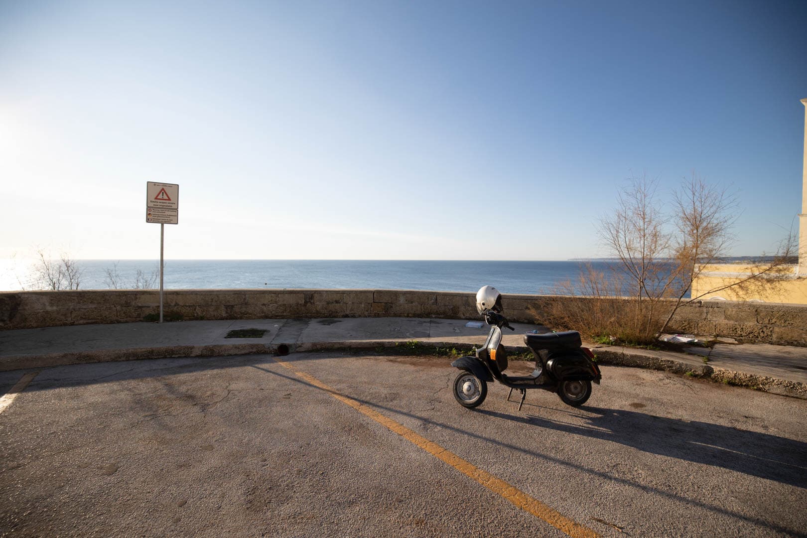 Ein schwarzer Motorroller mit einem weißen Helm auf dem Sitz ist auf einem leeren Parkplatz in der Nähe einer Steinmauer geparkt, von der aus man das Meer unter einem klaren blauen Himmel überblicken kann. Auf der linken Seite steht ein Straßenschild, und an der Mauer wachsen blattlose Sträucher. © Fotografie Tomas Rodriguez