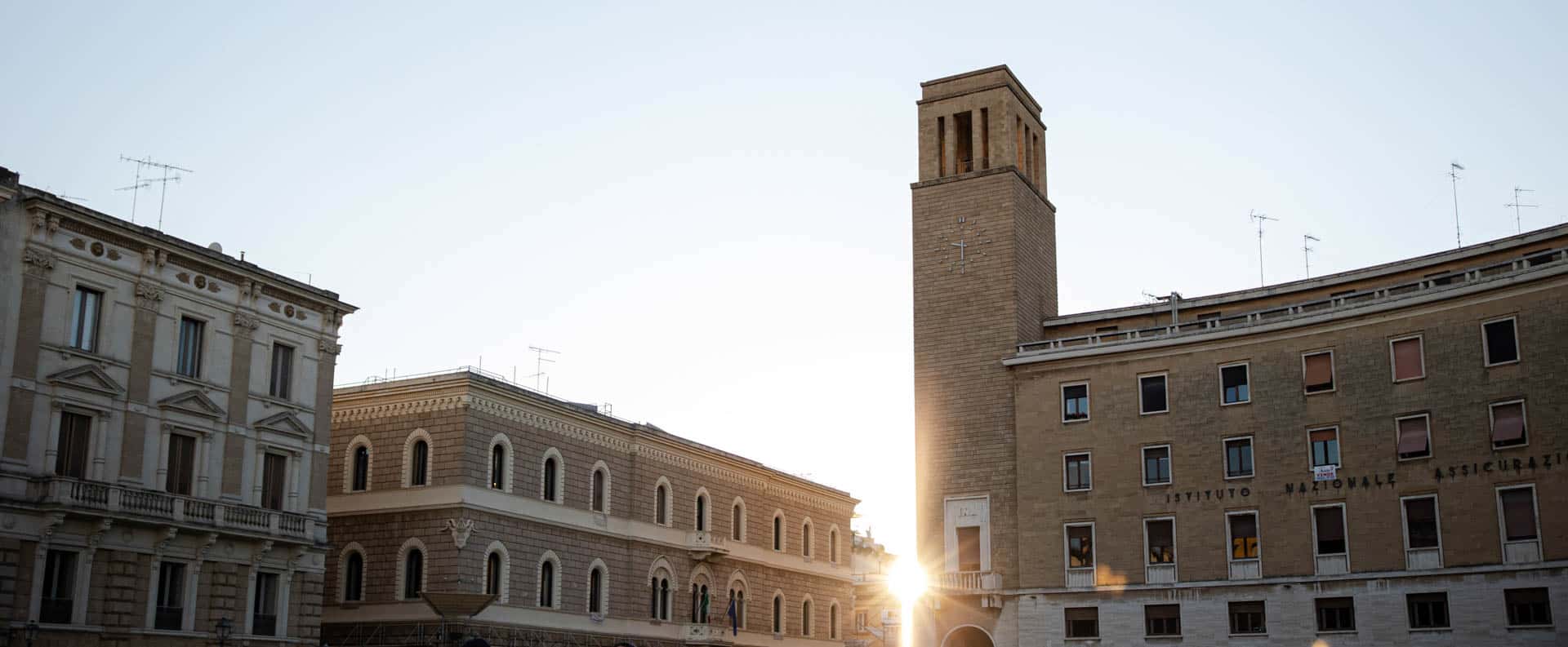 Sonnenaufgang durch historische Gebäude, darunter ein hoher Backsteinturm, auf einem italienischen Stadtplatz mit klassischer Architektur und klarem Himmel. © Fotografie Tomas Rodriguez