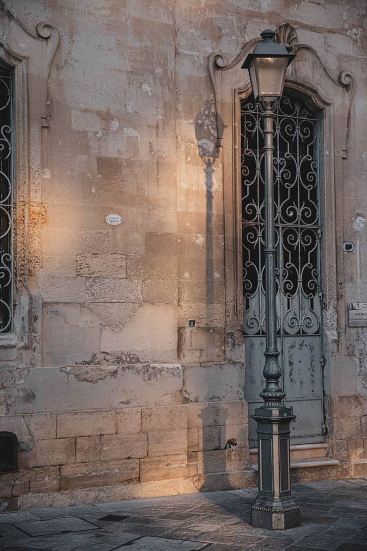 Eine alte Straßenlaterne steht vor einem alten Steingebäude mit verschnörkelten Eisentüren und verwitterten Wänden, das von warmem Sonnenlicht beleuchtet wird. Die Lampe wirft einen deutlichen Schatten auf die strukturierte Fassade. © Fotografie Tomas Rodriguez