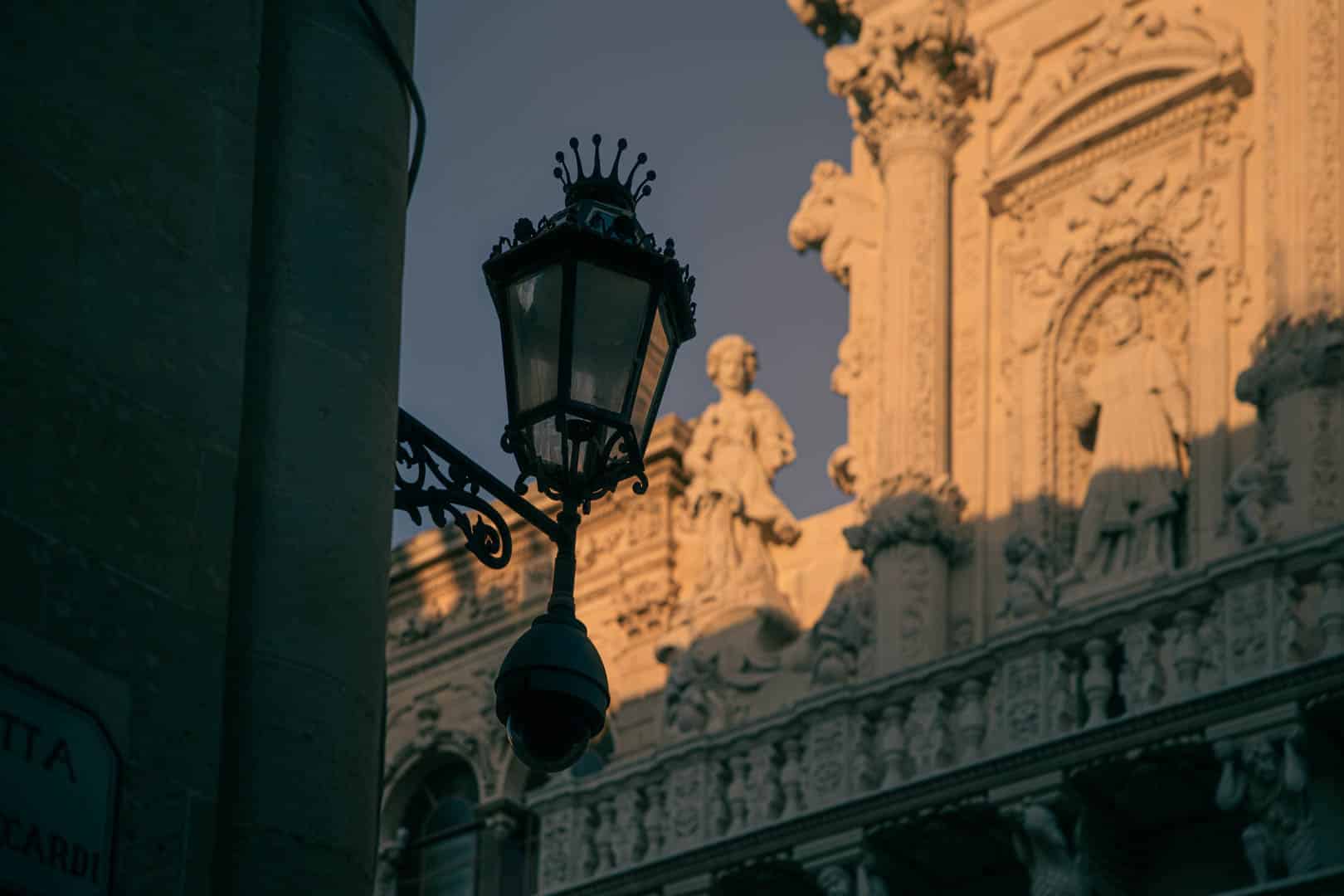 Eine verschnörkelte Straßenlaterne im Schatten ist neben einem historischen Gebäude mit kunstvollen Steinmetzarbeiten und Statuen zu sehen, das von warmem Sonnenlicht beleuchtet wird. Im Hintergrund sind detaillierte architektonische Reliefs und Figuren zu sehen. © Fotografie Tomas Rodriguez