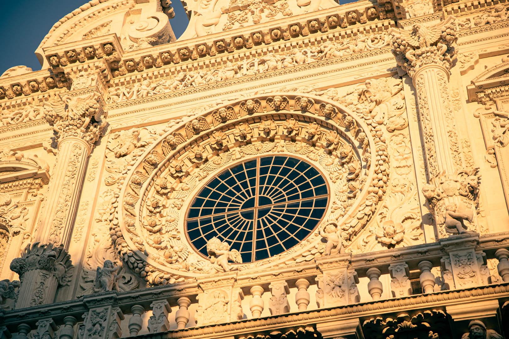 Detaillierte Nahaufnahme einer reich verzierten, sonnenbeschienenen Barockfassade mit kunstvollen Schnitzereien und einem großen runden Glasfenster in der Mitte, umgeben von geschnitzten Figuren und dekorativen Elementen. © Fotografie Tomas Rodriguez