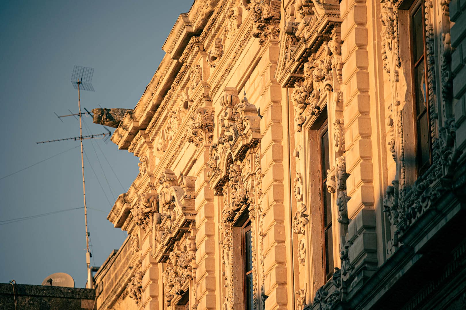 Nahaufnahme einer verzierten Gebäudefassade mit detaillierten Steinmetzarbeiten und Skulpturen, beleuchtet von warmem Sonnenlicht. Auf dem Dach ist eine Antenne vor einem klaren blauen Himmel zu sehen. © Fotografie Tomas Rodriguez