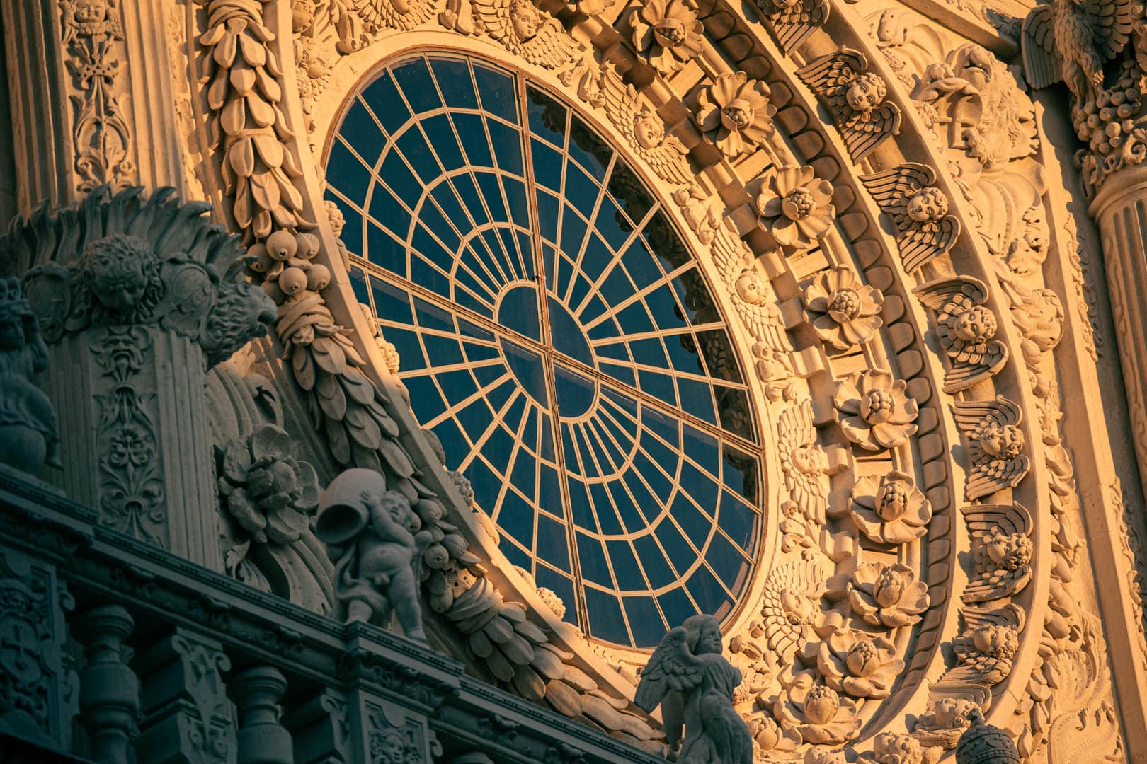 Nahaufnahme einer verschnörkelten Steinfassade mit einem großen, runden Buntglasfenster, umrahmt von kunstvollen floralen Schnitzereien und dekorativen Skulpturen, beleuchtet von warmem Sonnenlicht. © Fotografie Tomas Rodriguez