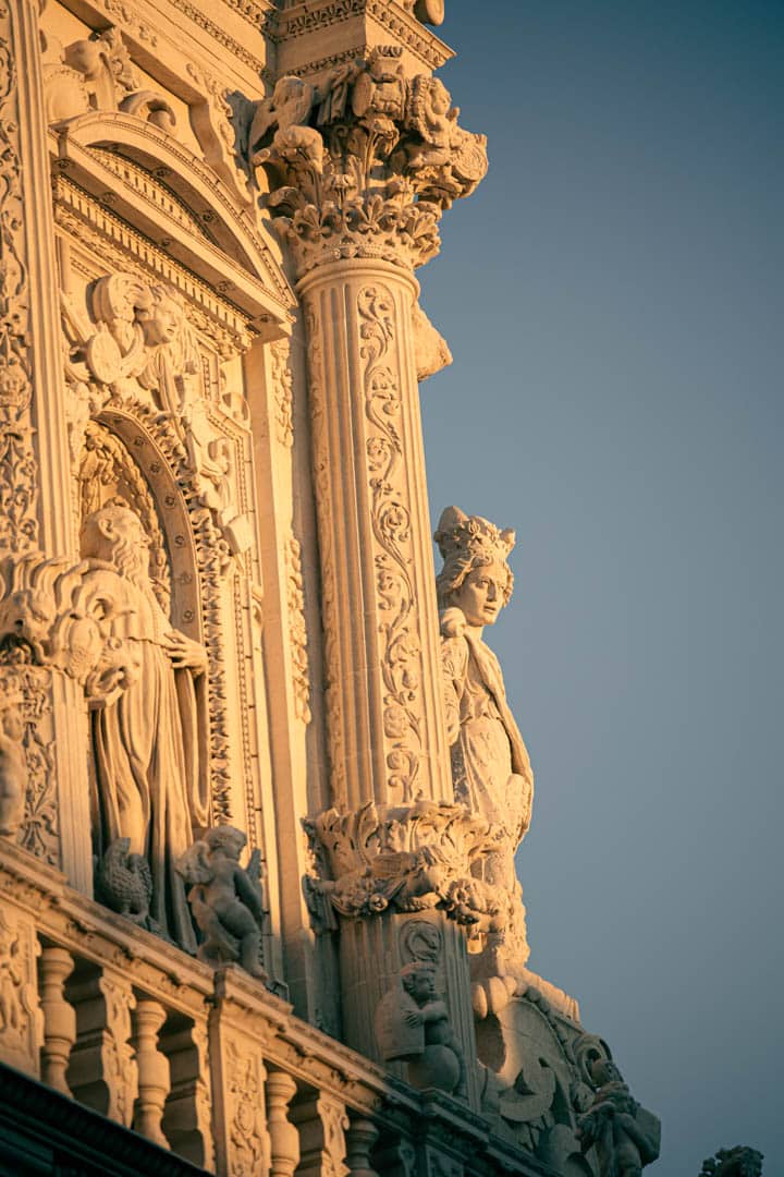 Verzierte Steinfassade in goldenem Sonnenlicht, mit detaillierten Schnitzereien, Säulen und einer prominenten Frauenfigur mit komplizierten Verzierungen vor einem klaren blauen Himmel. © Fotografie Tomas Rodriguez