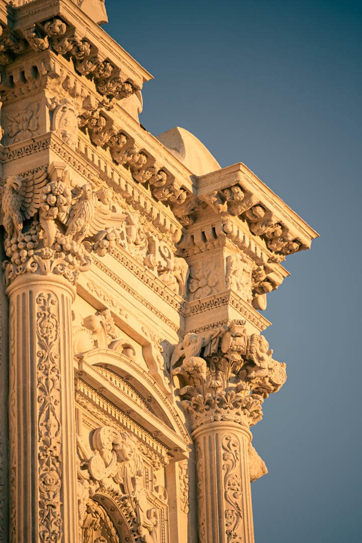 Detaillierte Nahaufnahme einer verschnörkelten, sonnenbeschienenen Steinfassade mit klassischen Säulen, Bögen und kunstvollen Schnitzereien von Blättern, Blumen und geflügelten Figuren vor einem klaren blauen Himmel. © Fotografie Tomas Rodriguez
