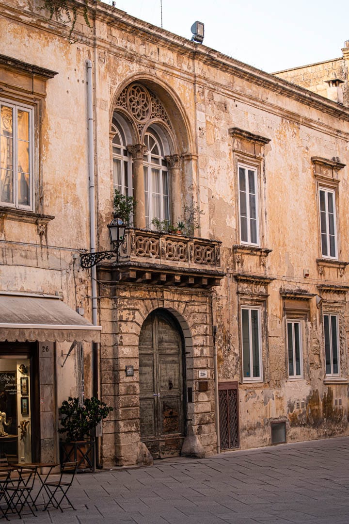 Ein altes, verwittertes Gebäude mit verschnörkelten Bogenfenstern, einer hölzernen Doppeltür und einem kleinen Balkon mit Topfpflanzen. Ein Café mit Tischen und Stühlen befindet sich neben dem Eingang an einer gepflasterten Straße. © Fotografie Tomas Rodriguez