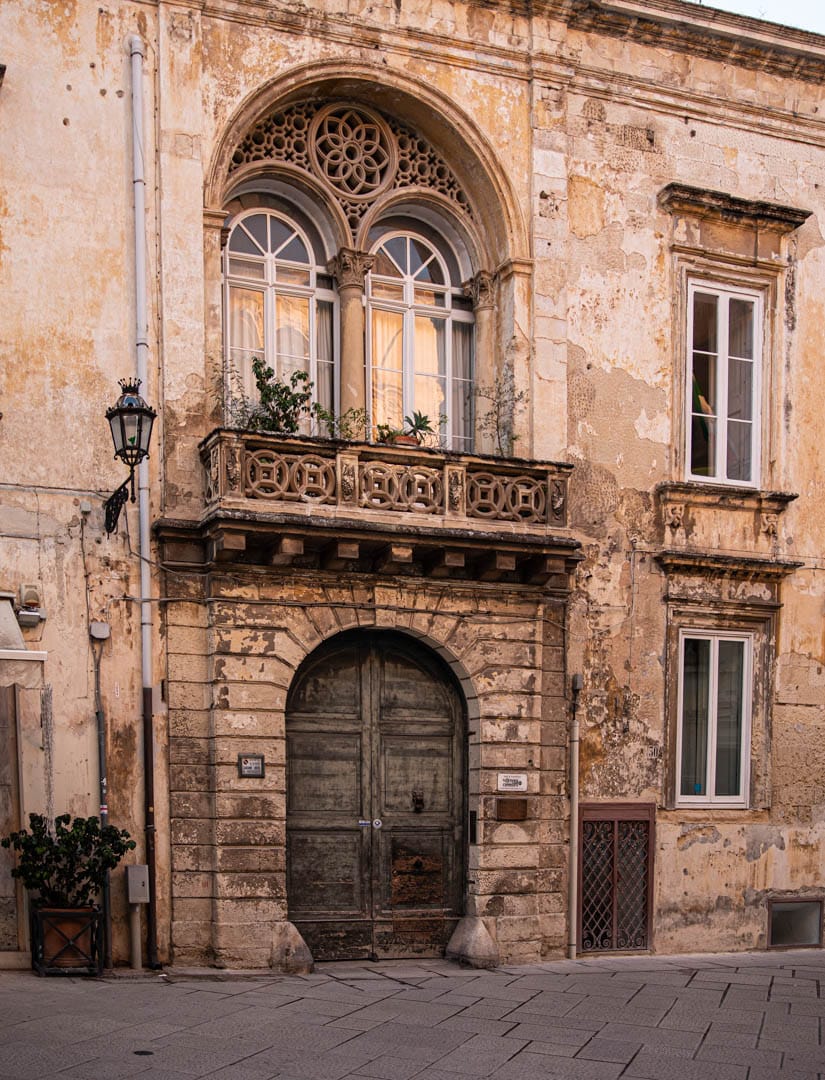 Ein altes, verwittertes Gebäude mit einer rustikalen, gewölbten Holztür, einem verschnörkelten Steinbalkon und zwei gewölbten Fenstern darüber. Die verblichenen beigen Wände zeigen Spuren des Alters, und auf dem Balkon stehen kleine Pflanzen. © Fotografie Tomas Rodriguez