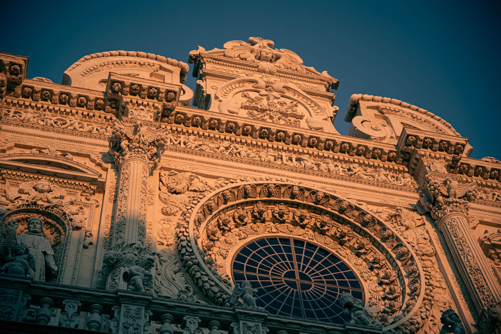 Verschnörkelte Barockfassade mit kunstvollen Schnitzereien, dekorativen Säulen und einem großen runden Buntglasfenster, das in warmes Sonnenlicht vor einem klaren blauen Himmel getaucht ist. © Fotografie Tomas Rodriguez
