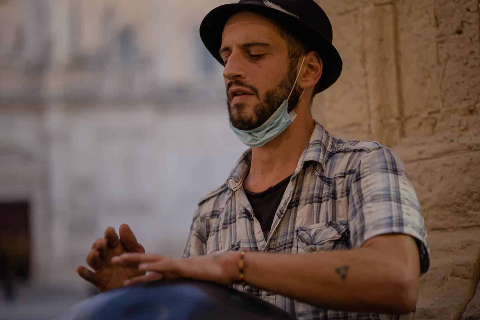 Ein Mann mit schwarzem Hut und kariertem Hemd spielt auf einer Handpan. Er hat einen Bart, eine unter das Kinn gezogene Gesichtsmaske und steht mit geschlossenen Augen an einer Steinmauer, die konzentriert zu sein scheint. © Fotografie Tomas Rodriguez