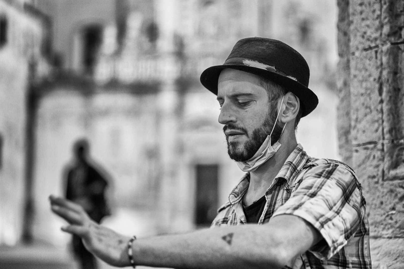 Ein Mann in einem karierten Hemd und einem Filzhut berührt eine Steinmauer und trägt eine Gesichtsmaske unter dem Kinn. Der Hintergrund ist unscharf und zeigt eine städtische Umgebung im Freien mit einer anderen Person, die unscharf ist. Das Bild ist in schwarz-weiß gehalten. © Fotografie Tomas Rodriguez