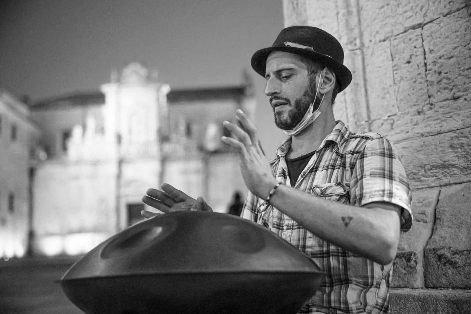 Ein Mann mit Hut und kariertem Hemd spielt nachts auf der Straße eine Handtrommel. Im Hintergrund ist ein Gebäude schwach beleuchtet, und unter seinem Kinn hängt eine Maske. Das Bild ist in schwarz-weiß gehalten. © Fotografie Tomas Rodriguez