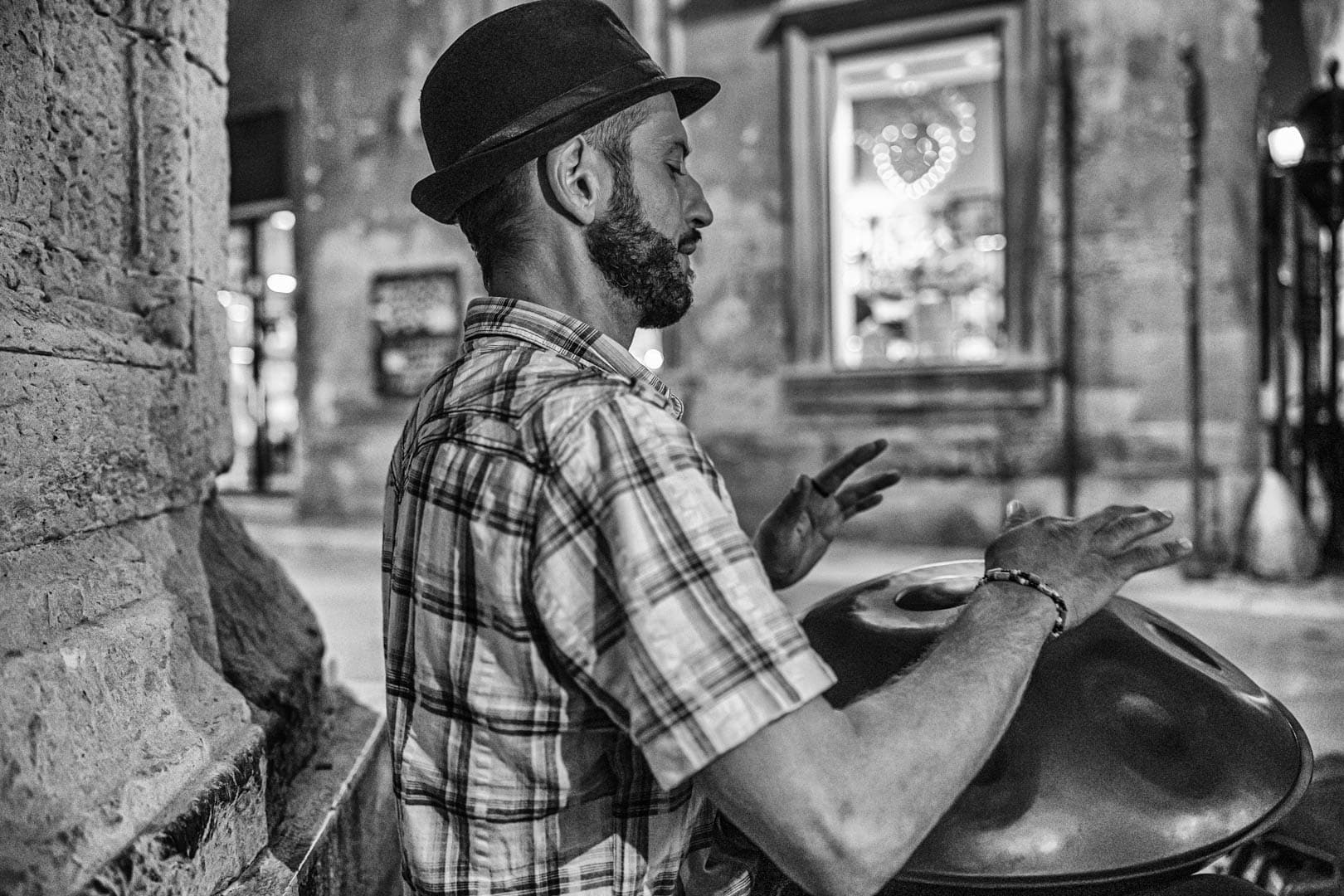 Ein Mann mit kariertem Hemd und Hut spielt auf einer nächtlichen Straße in der Stadt eine Handtrommel. Die Szene ist in Schwarz-Weiß gehalten, mit unscharfen Gebäuden und einem Schaufenster im Hintergrund. © Fotografie Tomas Rodriguez