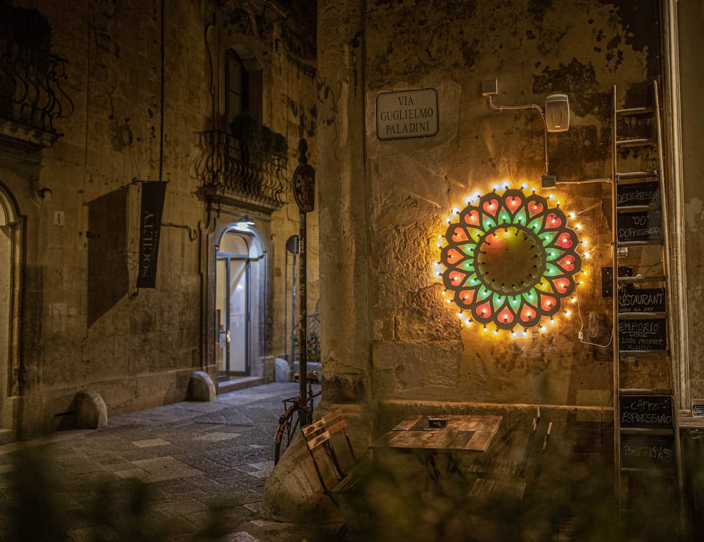 Eine warm beleuchtete Straßenecke in der Nacht zeigt eine bunte, kreisförmige Lichtdekoration an einer Steinmauer unter dem Schild "Via Guglielmo Paladini". In der Nähe stehen ein Holztisch und Stühle, die engen Gassen und Balkone sind zu sehen. © Fotografie Tomas Rodriguez