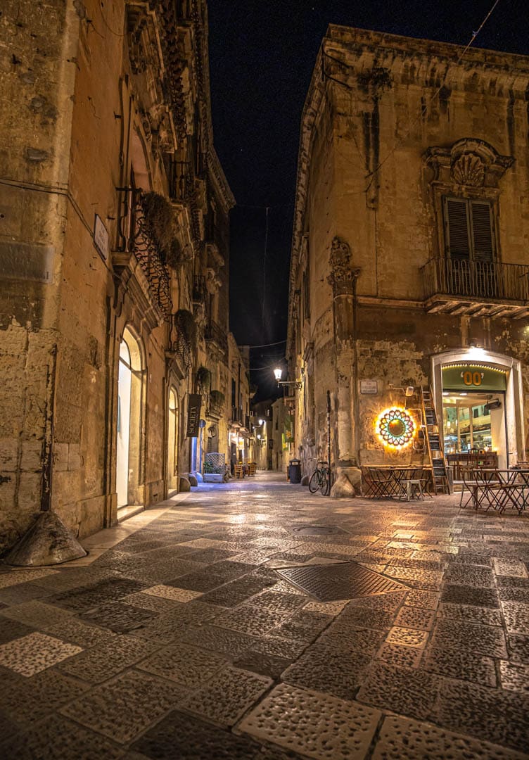 Eine schmale, kopfsteingepflasterte Straße bei Nacht in einer alten europäischen Stadt, mit warm beleuchteten Geschäften und abgenutzten Steinhäusern auf beiden Seiten. Vor einem der Läden stehen ein paar Tische und ein leuchtendes Schild. © Fotografie Tomas Rodriguez